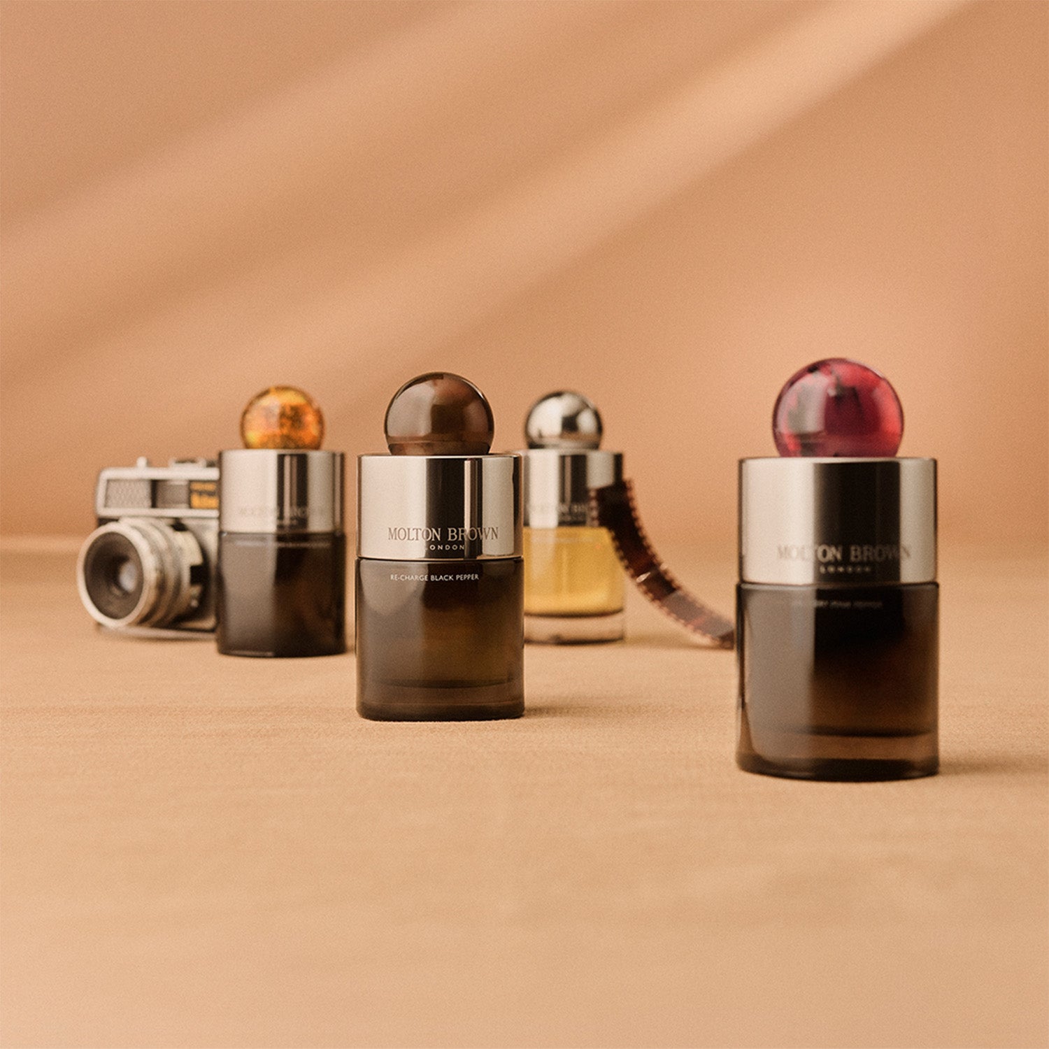 Four Molton Brown perfume bottles are arranged on a beige surface, with an old-fashioned camera and film strip in the background. Each bottle has a colorful, spherical cap.
