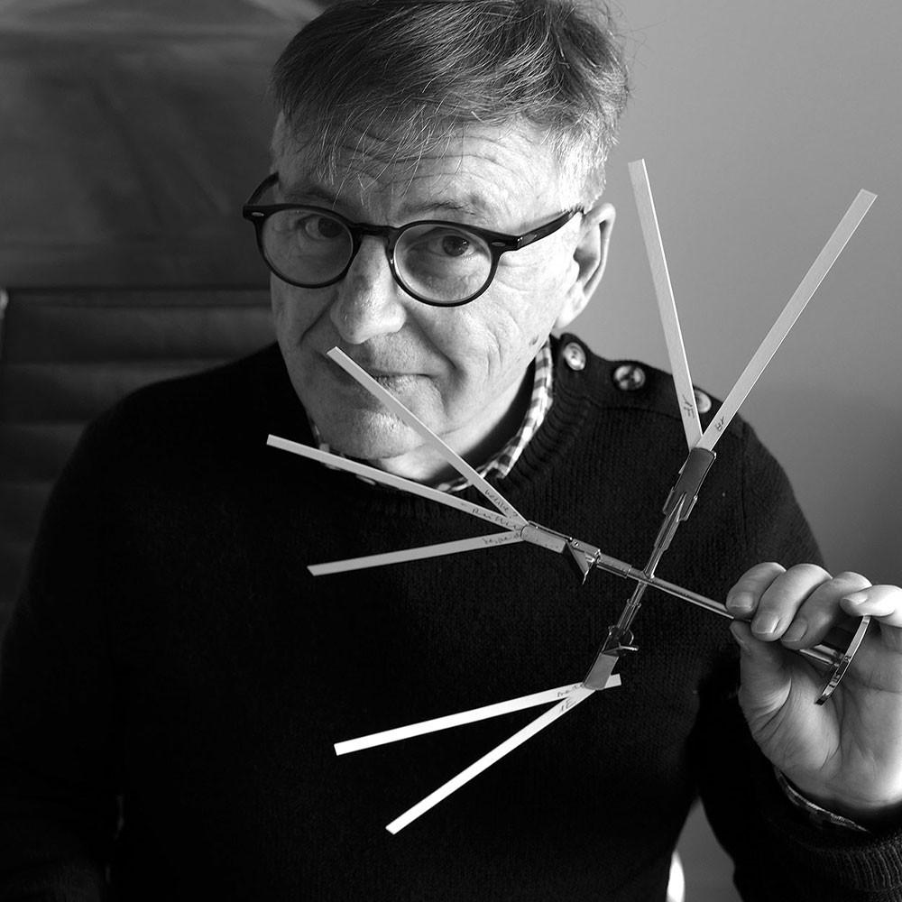 A man wearing glasses and a dark sweater holds a geometric tool with multiple extending arms, looking at the camera. The photo is in black and white.