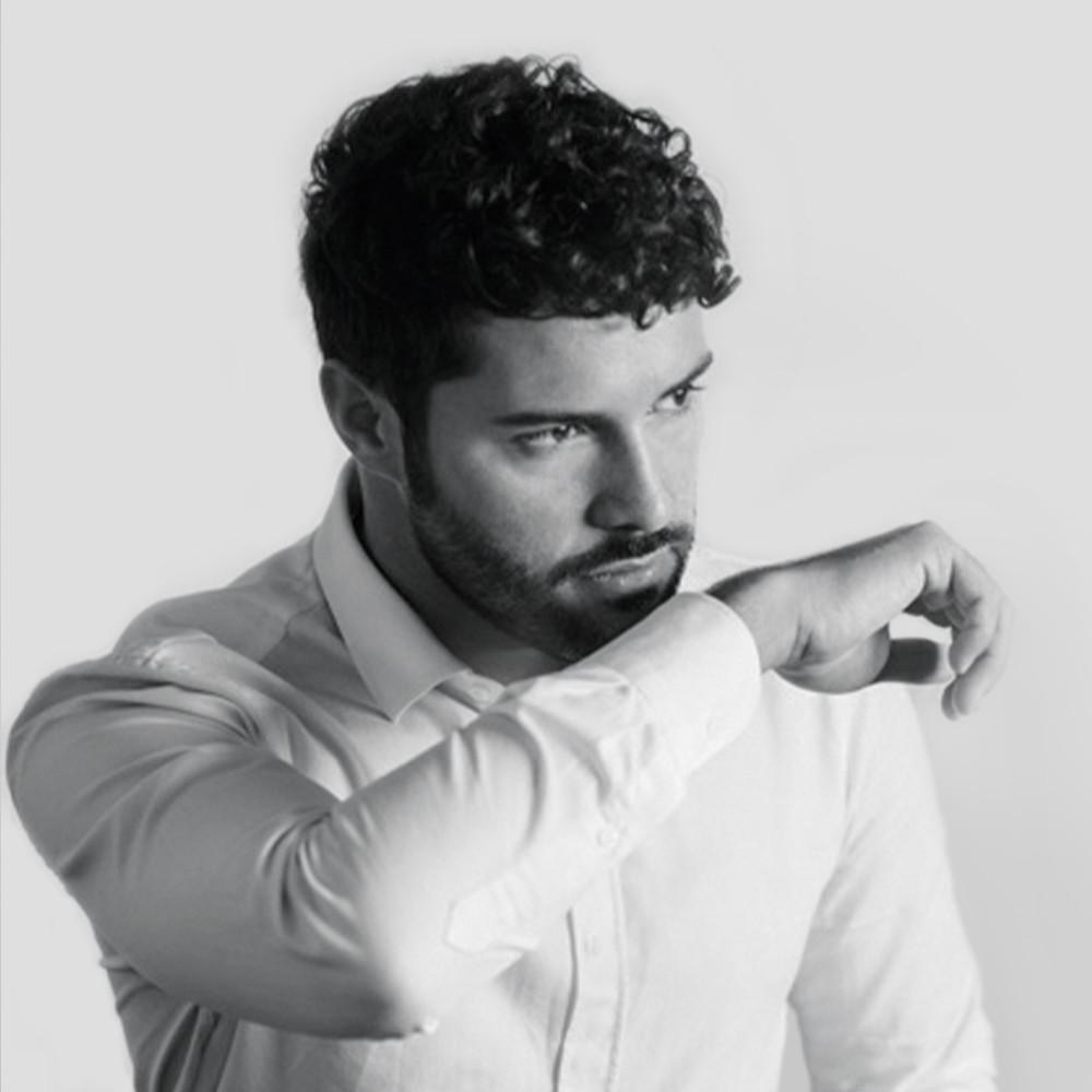 A man with curly dark hair and a beard, wearing a light-colored button-up shirt, looks to the side while resting his forearm against his mouth. The background is plain and light.