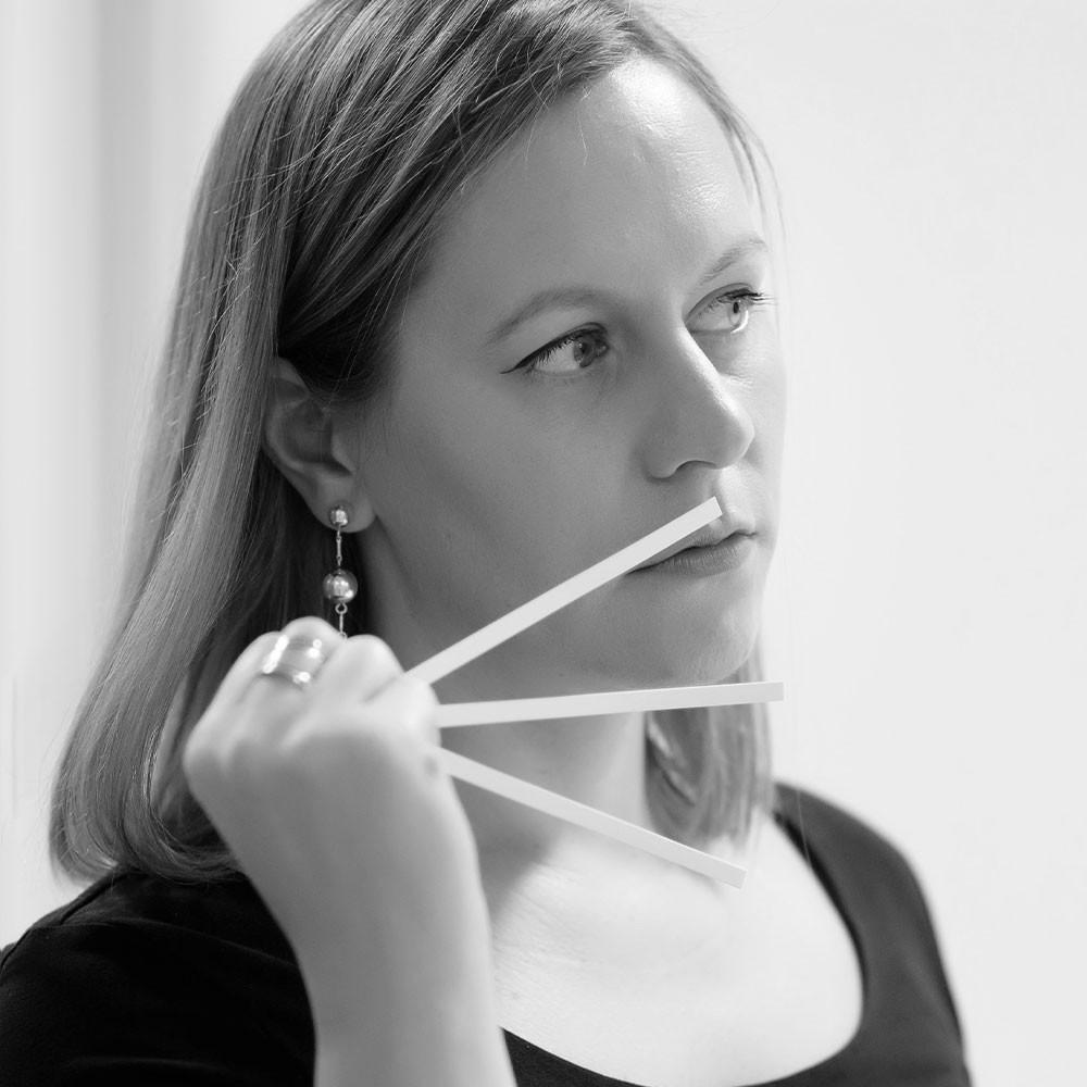 A woman with straight, shoulder-length hair holds three scent testing strips near her nose, appearing thoughtful. She wears a dark top and drop earrings. The photo is in black and white.
