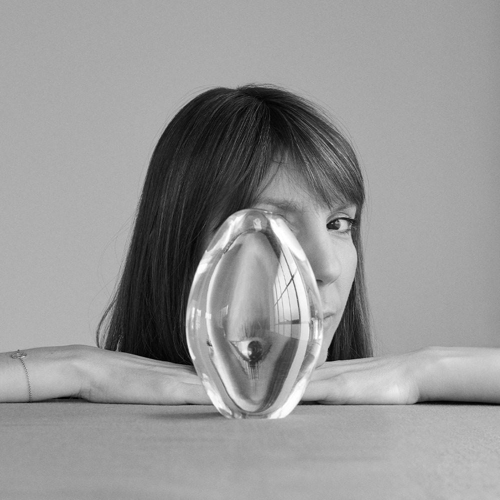 A woman rests her chin on her hands atop a table, gazing through a large, oval glass object that distorts the appearance of one of her eyes. The image is in black and white.