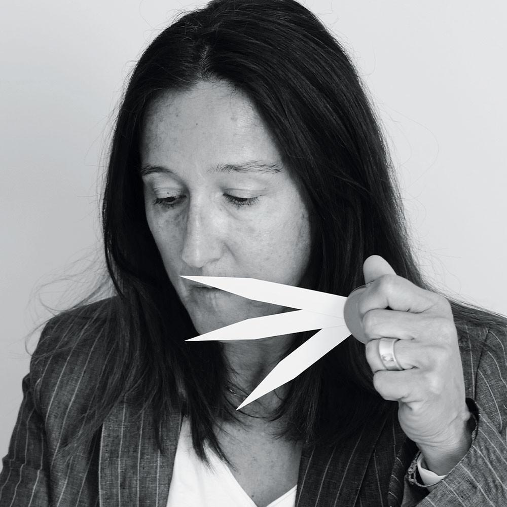 A woman with long dark hair holds up three white paper strips near her face, appearing to smell them. She is wearing a striped blazer and looking down, with a neutral background behind her.