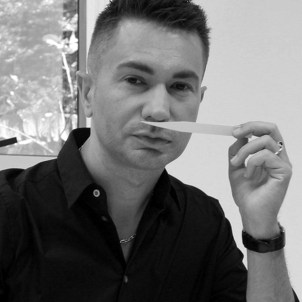A man wearing a dark shirt holds a scent testing strip under his nose, appearing to evaluate a fragrance. He is indoors, with a window showing trees in the background. The image is in black and white.