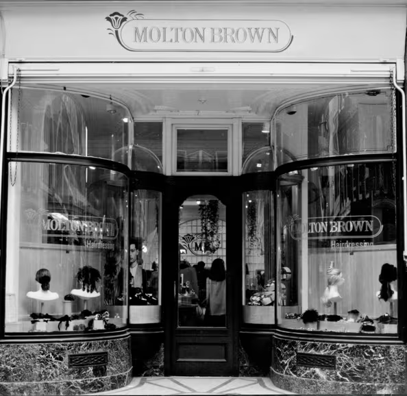 Black and white photo of a Molton Brown storefront with large curved display windows showing wigs on stands and people visible inside the shop.