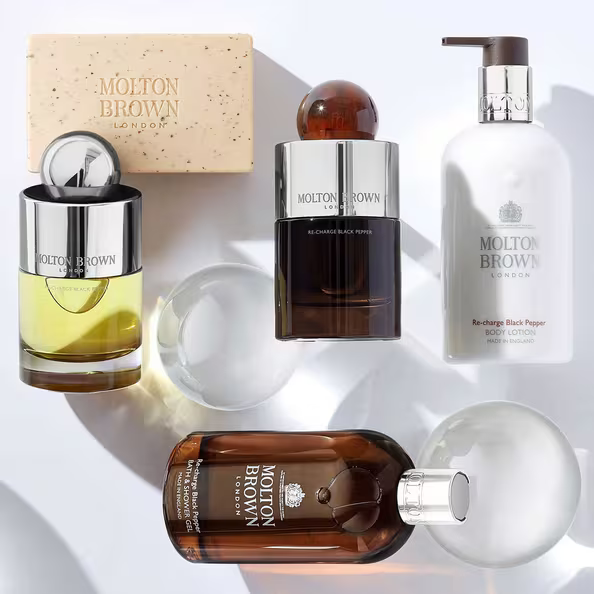 Molton Brown products, including soap, cologne, body wash, and lotion, are arranged on a white surface with two clear glass spheres casting soft shadows.