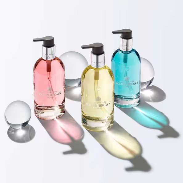 Three Molton Brown soap bottles in pink, yellow, and blue with pump tops are placed on a white surface, casting colored shadows. Two clear glass spheres sit next to them, adding reflections and depth.