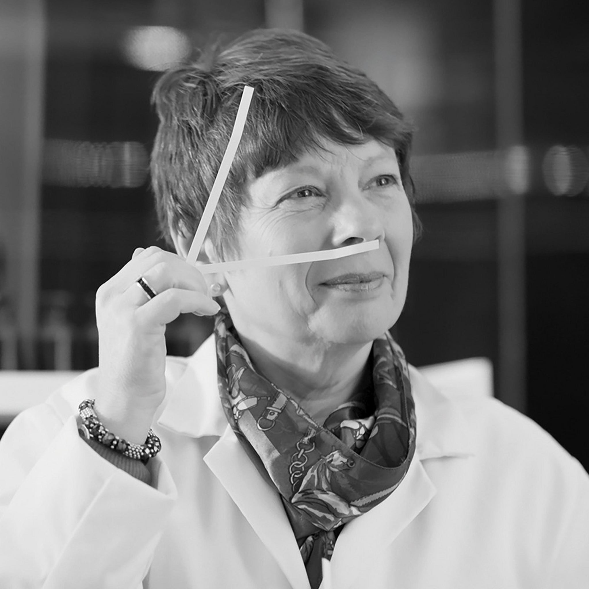 A woman in a lab coat and scarf holds two scent strips under her nose, appearing to evaluate their smell, in a laboratory setting.