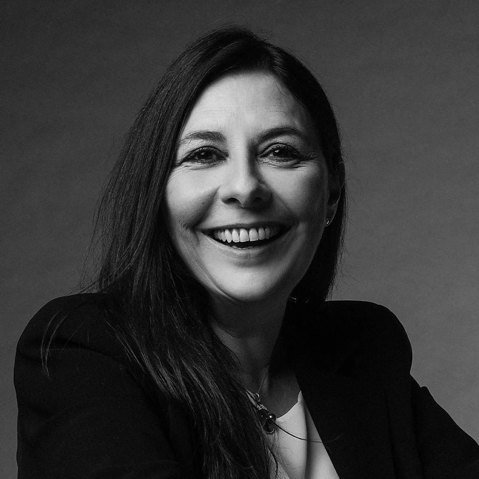 Black and white portrait of a woman with long dark hair, smiling warmly at the camera. She is wearing a blazer and a light top, posed against a plain background.