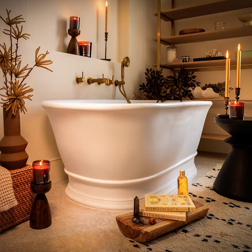 A cozy bathroom with a white freestanding tub, gold fixtures, candles, dried plants, and books on a wooden tray, creating a warm, relaxing atmosphere. Shelves with towels and decor are in the background.