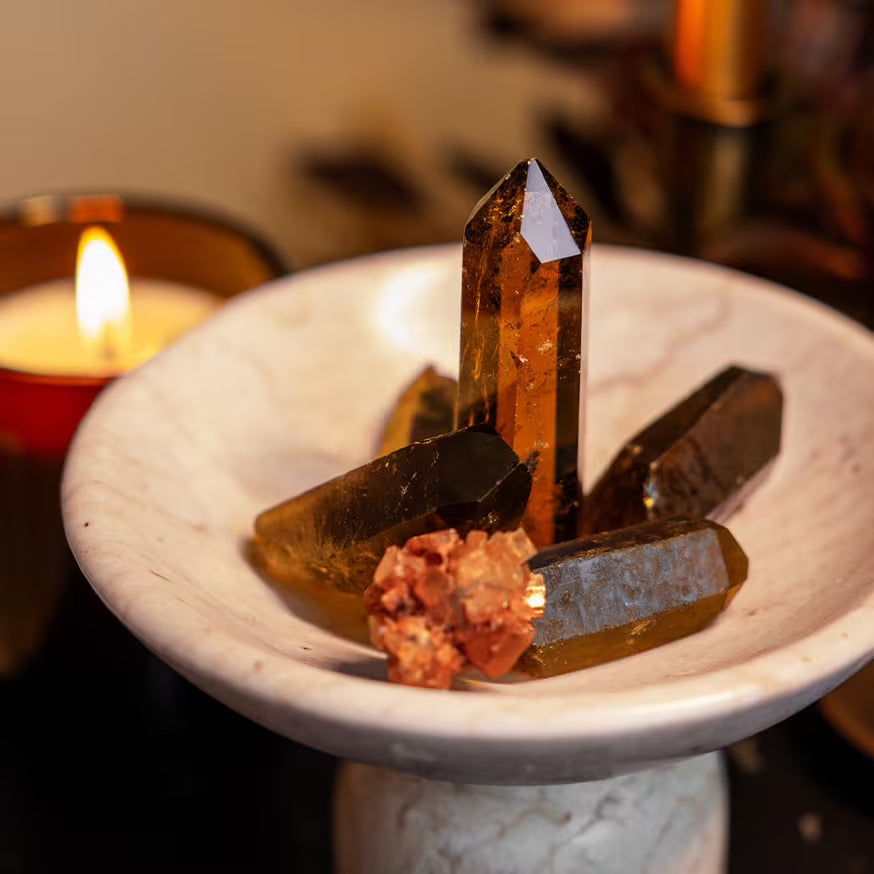 A white marble dish holds several polished brown crystals and a cluster of red stones, with a lit candle in the background creating a warm, ambient glow.