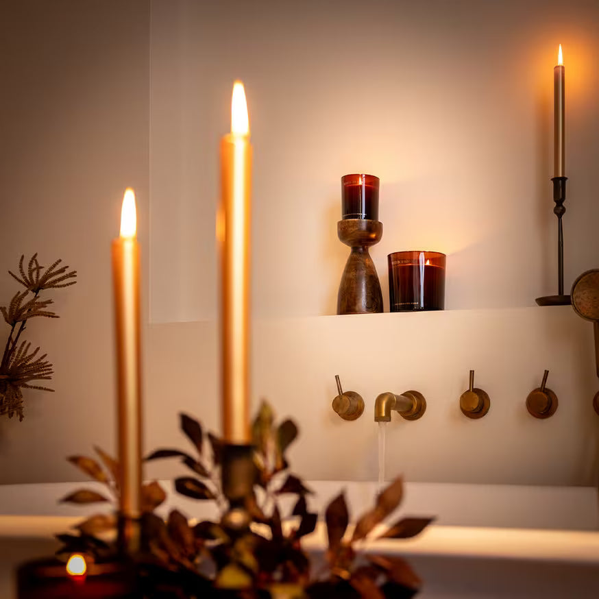 A cozy bathroom scene with a white tub, lit candles in holders, bronze taps, and leafy decor, creating a warm, relaxing atmosphere with soft lighting.
