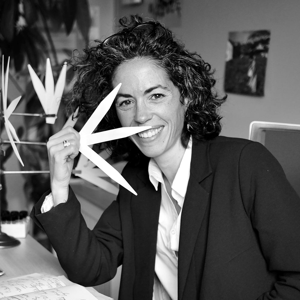 A woman with curly hair, wearing a blazer, sits at a desk and smiles at the camera while holding a large, stylized leaf shape up to her face in an office setting.