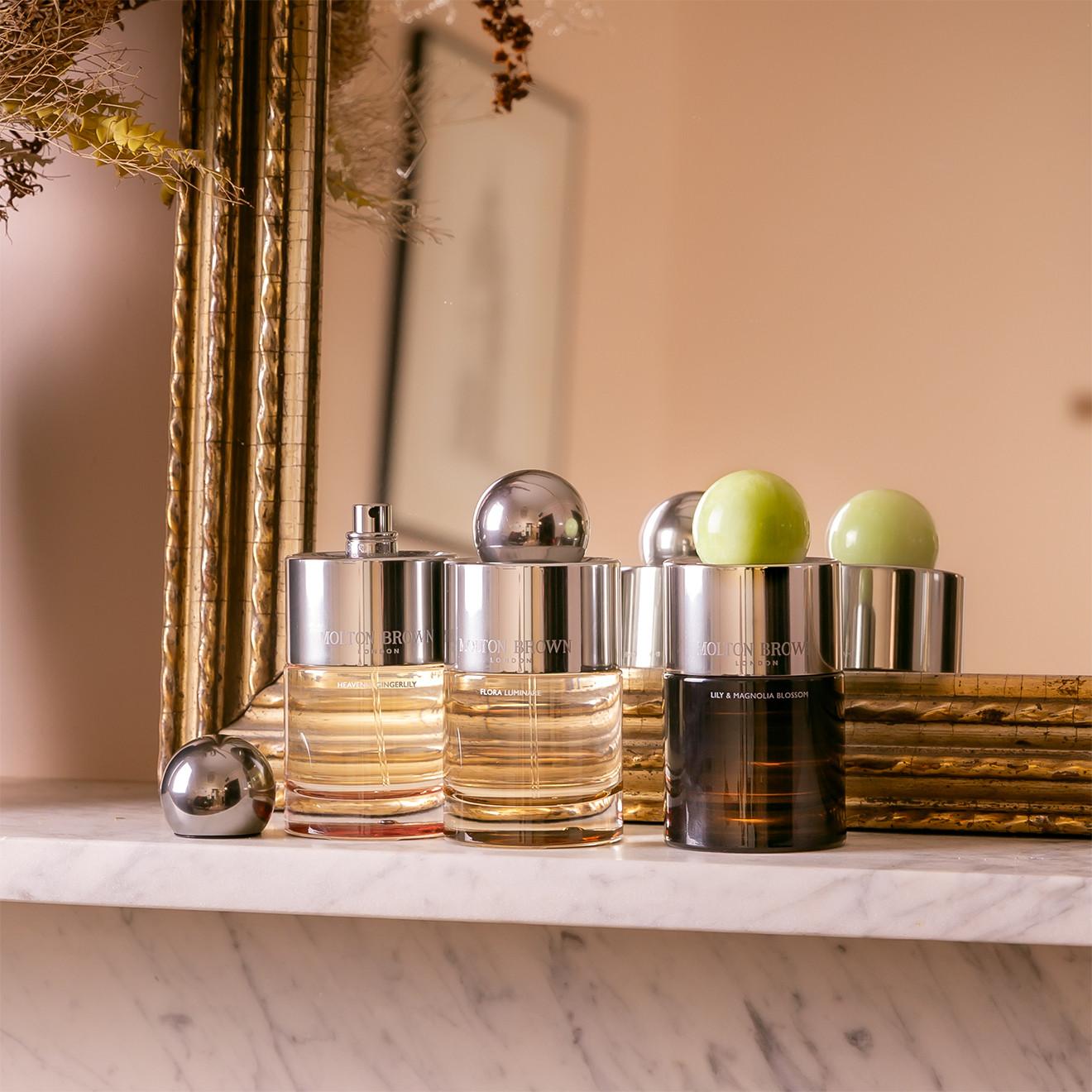 Three perfume bottles with metallic caps are arranged on a marble surface in front of a gold-framed mirror. Two bottles have silver caps and light amber liquid, while the third has a green, round cap and dark liquid. Dried flowers hang above.
