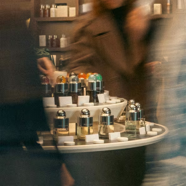 A circular display of assorted perfume bottles in a store, surrounded by blurred figures of people moving past.