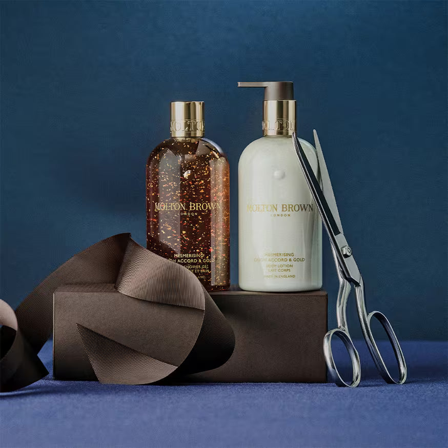 Two Molton Brown bottles, one with a pump and one with a screw cap, sit on a black box next to large silver scissors and a black ribbon, all set against a dark blue background.