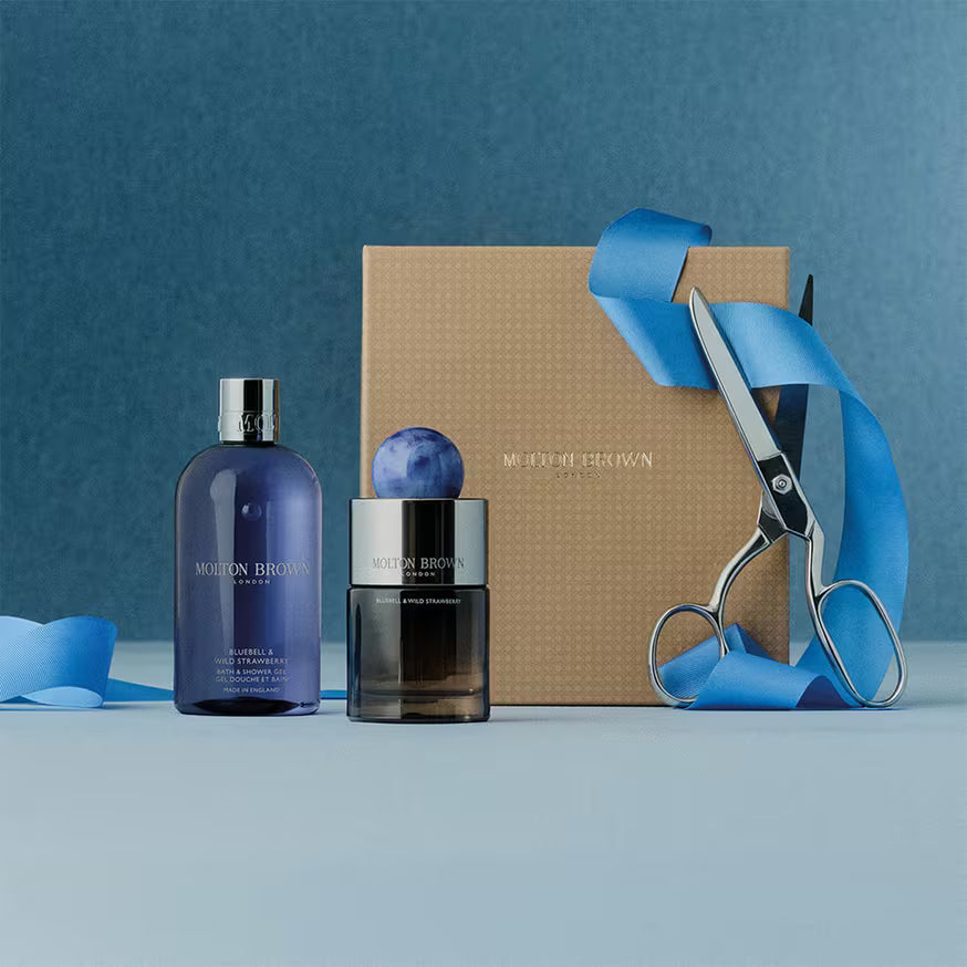 A Molton Brown gift set with a gold box, a blue bottle, a jar with a blue marble lid, scissors, and blue ribbon, arranged on a blue surface against a blue background.