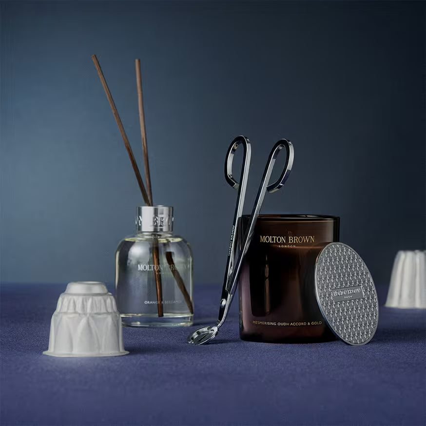 A Molton Brown candle with a silver lid, candle wick trimmer, and a glass reed diffuser sit on a blue surface alongside two white decorative objects. The background is also blue.