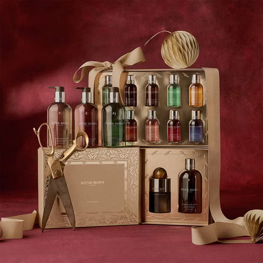 A luxury Molton Brown gift set on a red background, featuring various scented bottles, a gold embossed box, large gold scissors, and ribbon, with decorative paper ornaments.