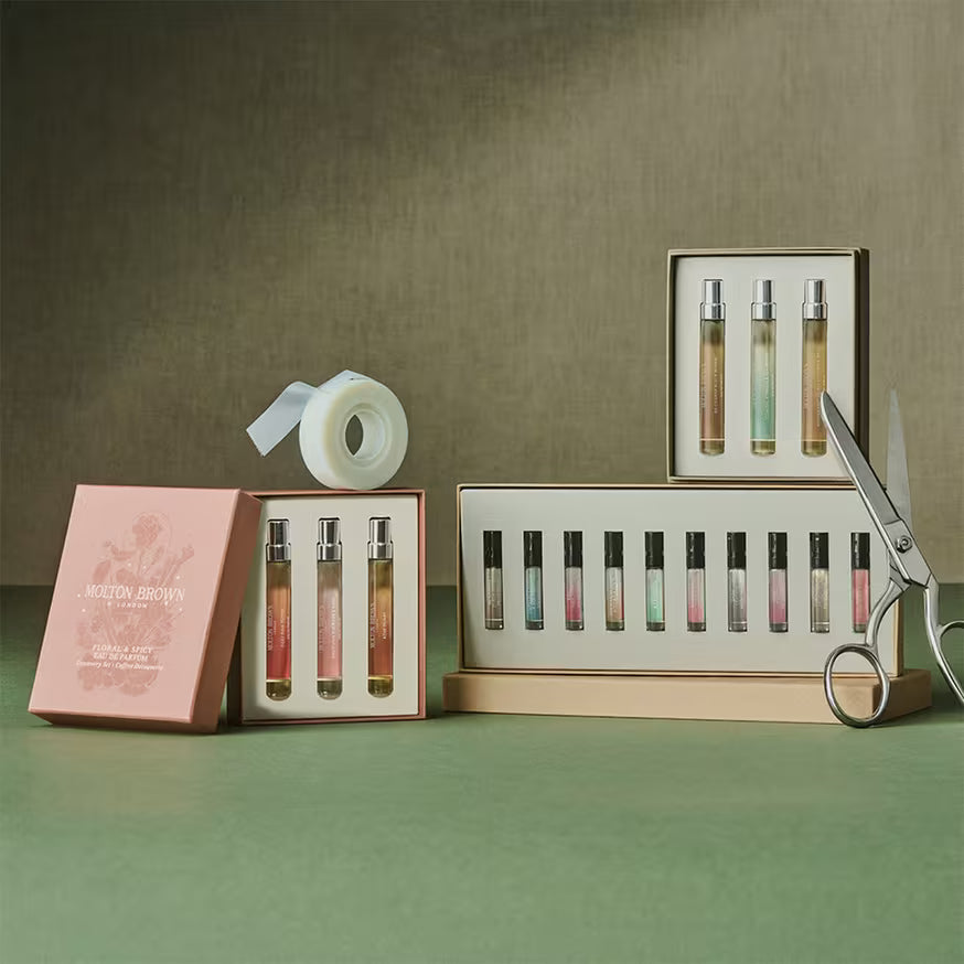 A display of Molton Brown fragrance gift sets with small perfume vials, an open pink box, a roll of white ribbon, and a pair of scissors on a green surface against a muted background.