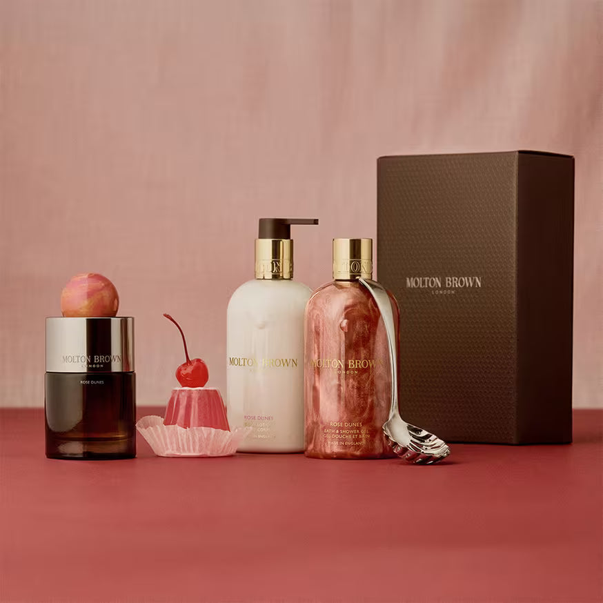 Molton Brown gift set with a candle, lotion, and body wash in elegant packaging, displayed with a decorative cherry and a spoon on a red surface with a pink background.