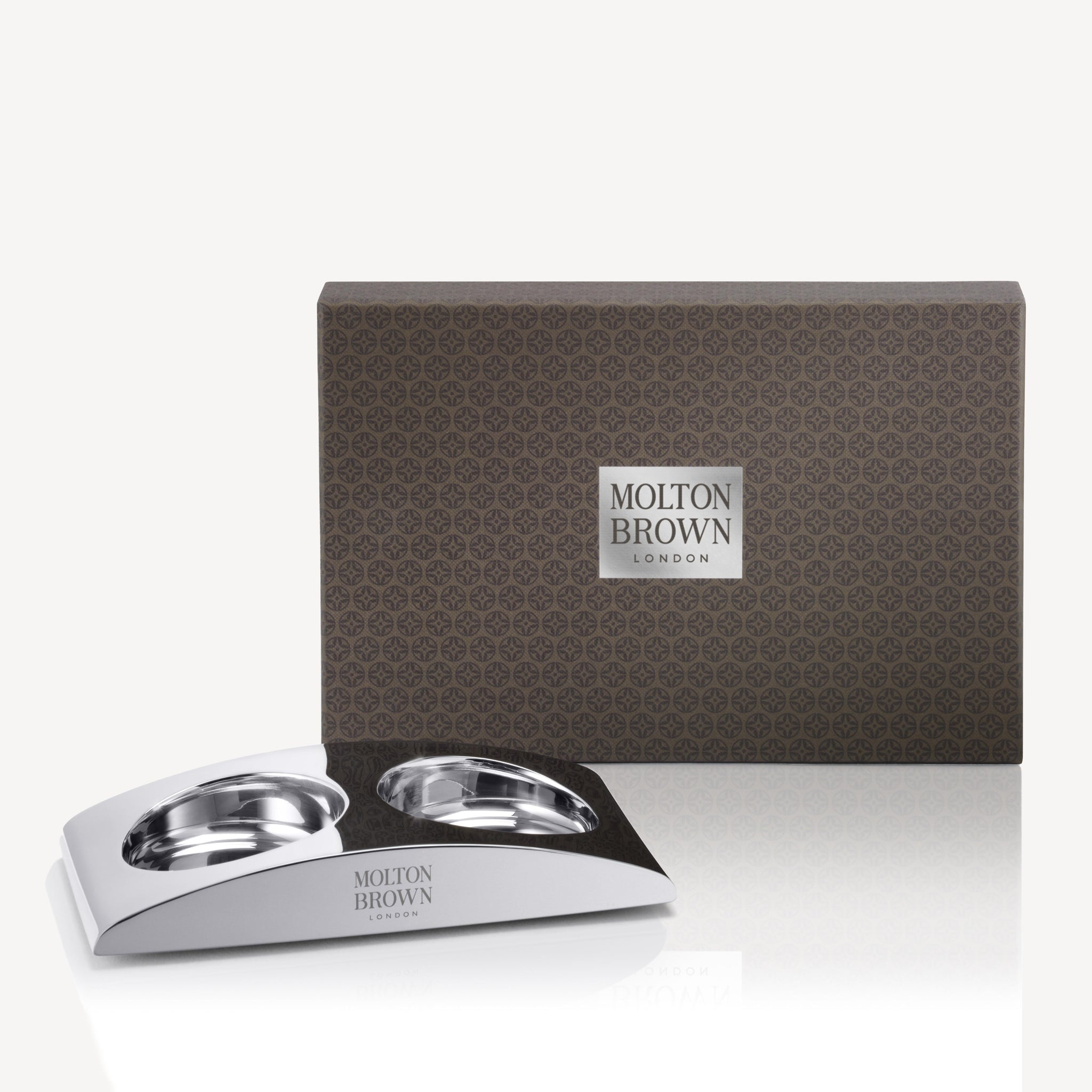 A rectangular brown MoltonBrown Nederlands The Elemental Arc gift box stands behind a sleek silver lotion holder with two round compartments, both featuring the MoltonBrown Nederlands logo.