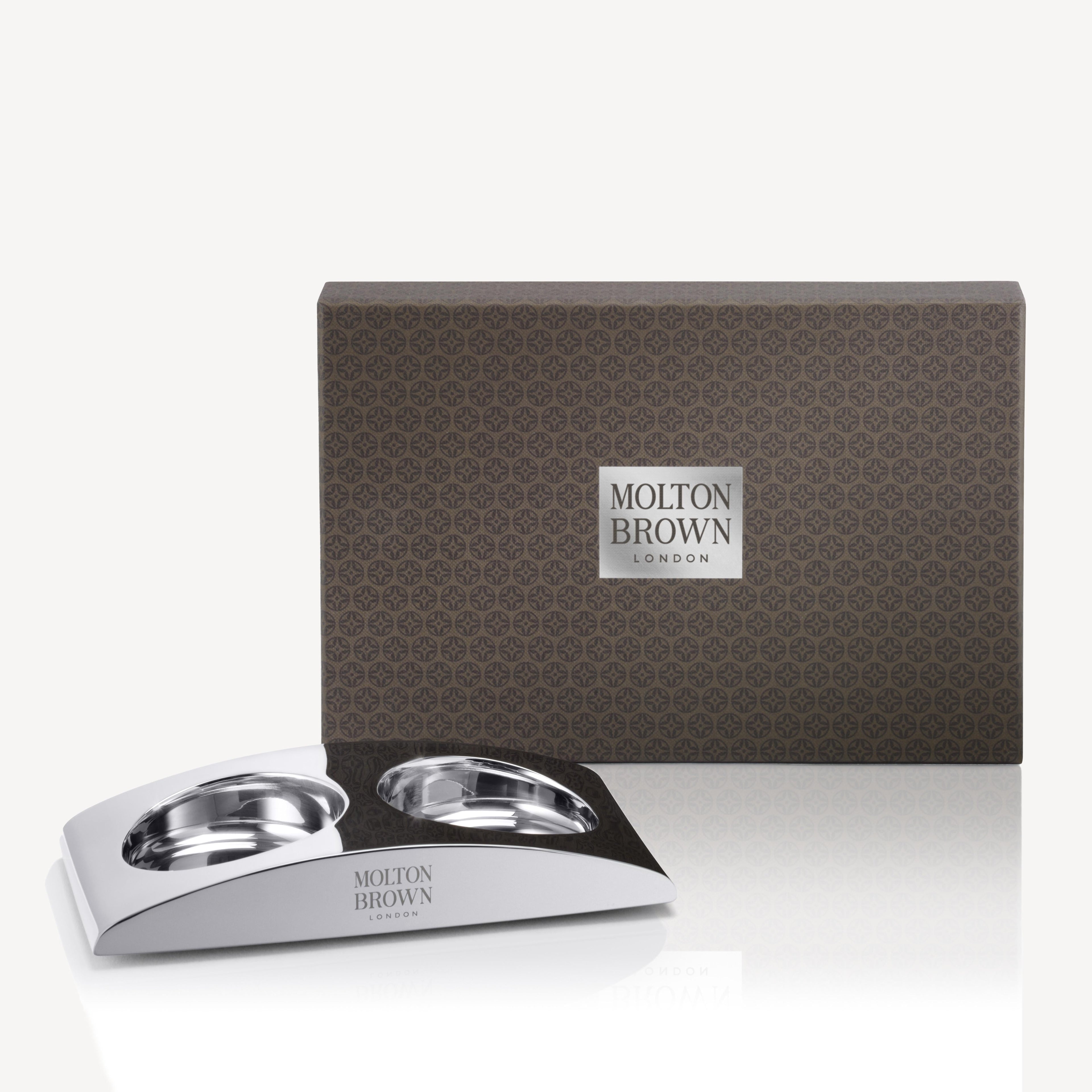 A rectangular brown MoltonBrown Nederlands The Elemental Arc gift box stands behind a sleek silver lotion holder with two round compartments, both featuring the MoltonBrown Nederlands logo.