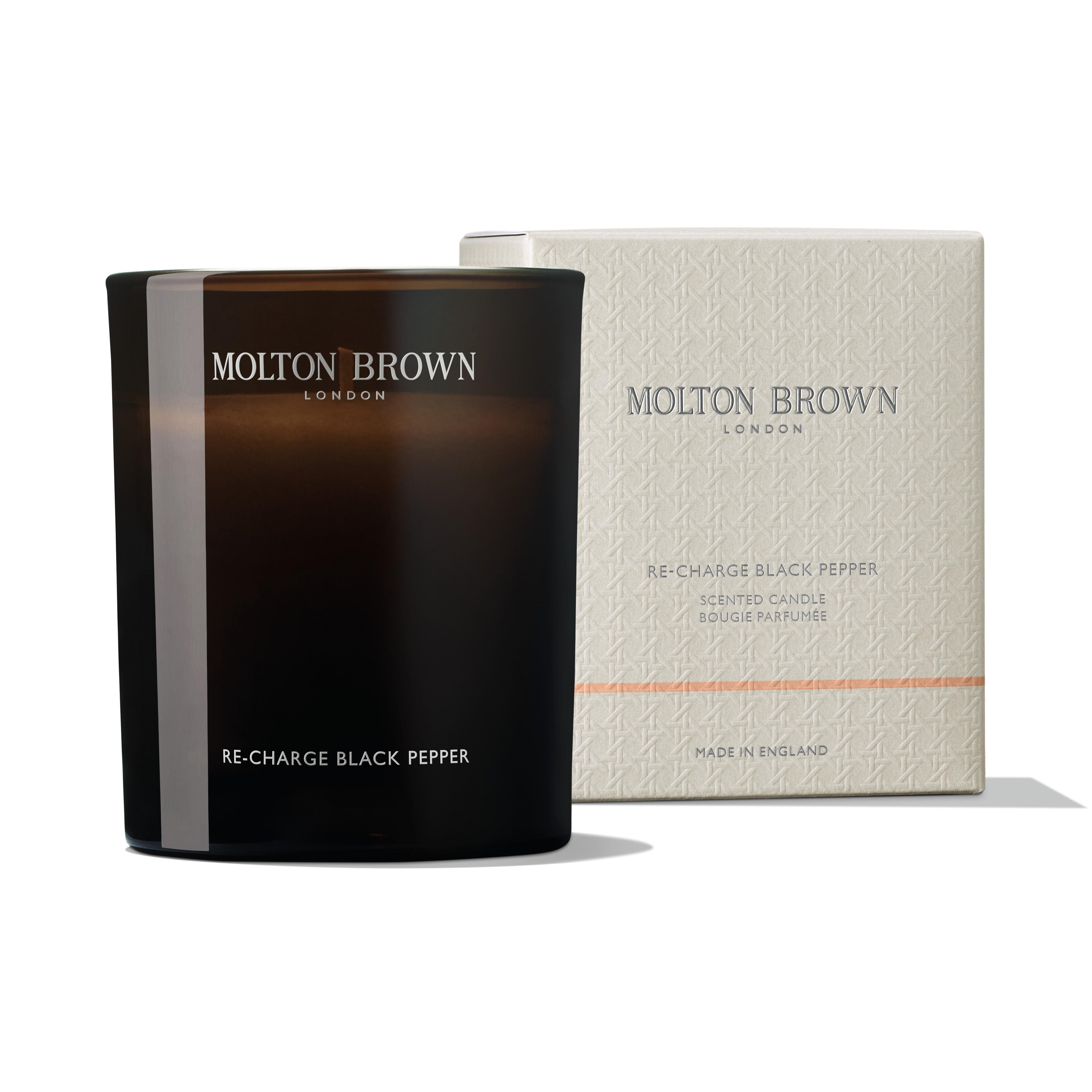 The MoltonBrown Nederlands Re-charge Black Pepper Signature Kaars 190g in een donker glazen houder staat voor zijn crèmekleurige doos met zilveren en oranje accenten, en verspreidt frisse citrus- en houtachtige tonen.