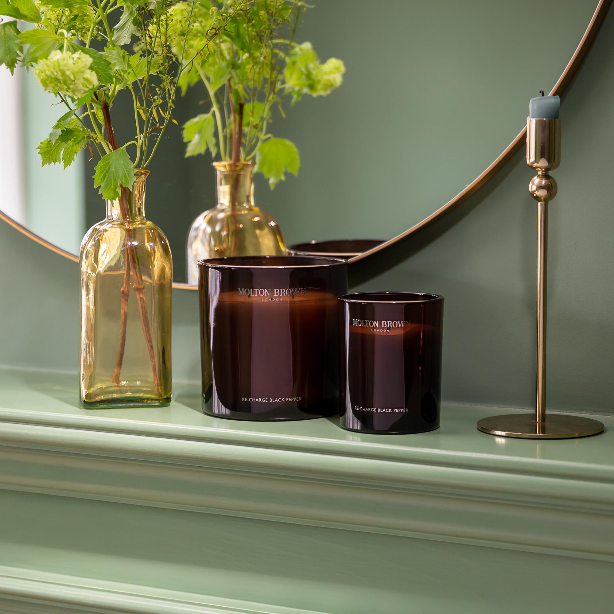 Two dark brown MoltonBrown Nederlands Re-charge Black Pepper Signature Kaars 190g candles rest on a light green shelf next to a gold vase with green foliage and a gold holder with a blue candle, all mirrored in a round reflection.