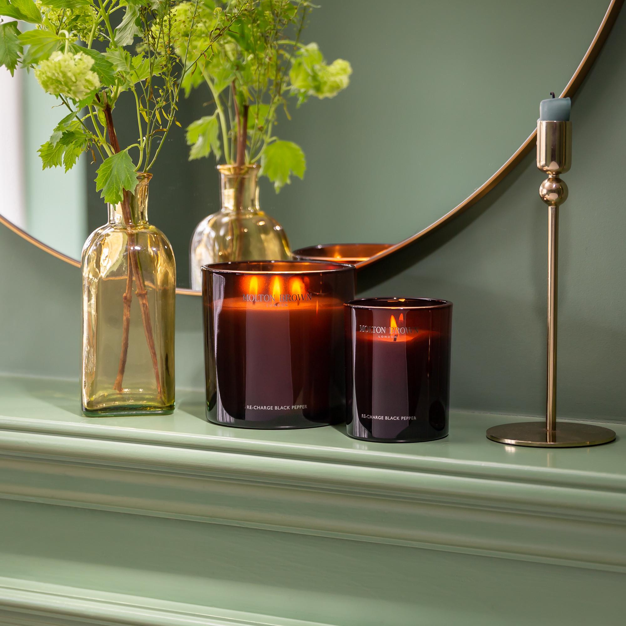Two lit dark glass candles, including the MoltonBrown Nederlands Re-charge Black Pepper Signature Kaars 190g with houtachtige tonen en frisse citrus, sit on a light green mantel beside a brass vase of leafy stems, round mirror, and brass candlestick with blue candle.