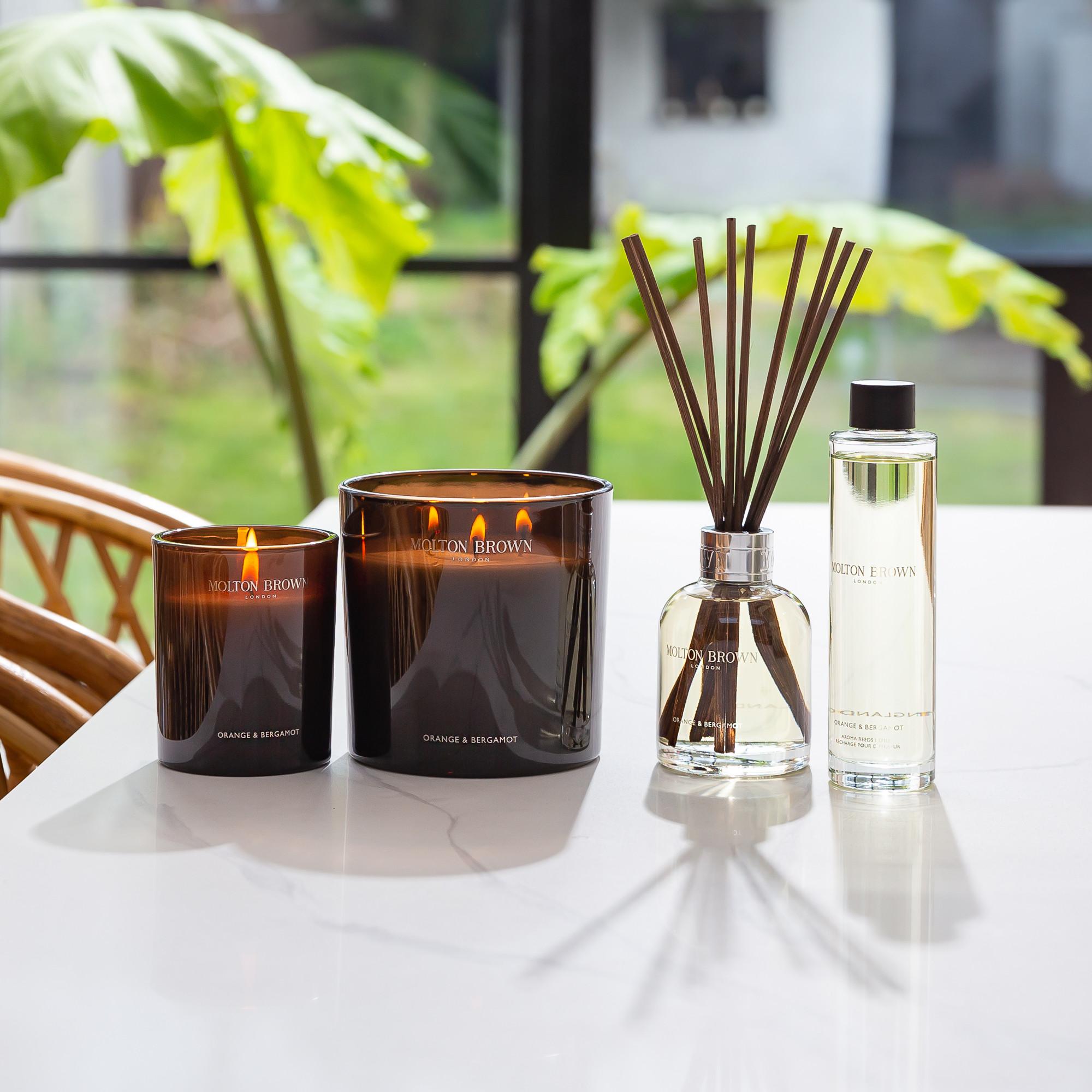 Two lit MoltonBrown Nederlands Orange & Bergamot Luxury Kaars 600g candles, a clear glass diffuser with reeds, and a matching bottle sit on a white tabletop in a bright room, infusing the air with citrus fragrance.