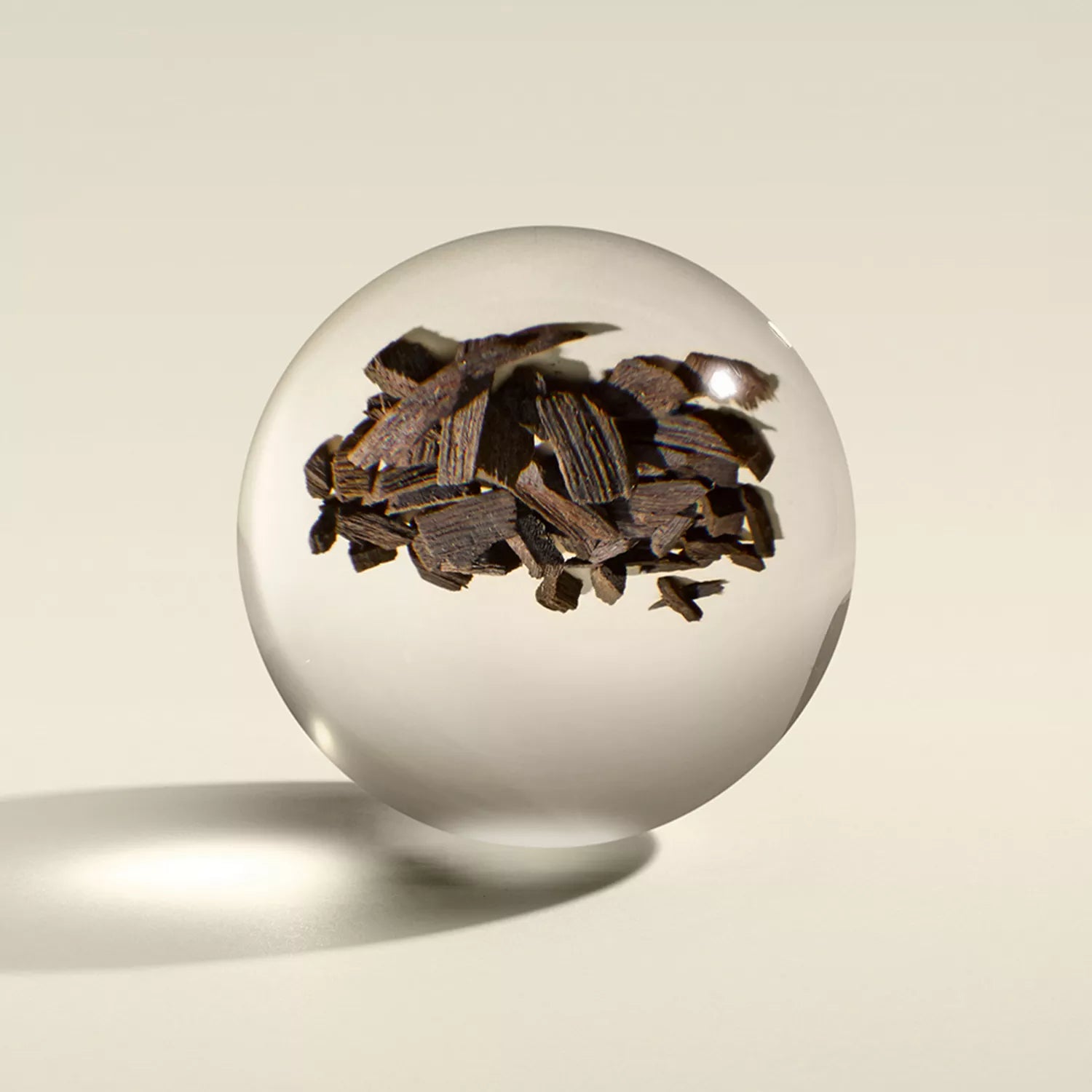 A clear glass sphere floats above a light background, with dark brown wood chips suspended inside—evoking the rich notes of Mesmerising Oudh Accord & Gold Signature Kaars 190g by MoltonBrown Nederlands.