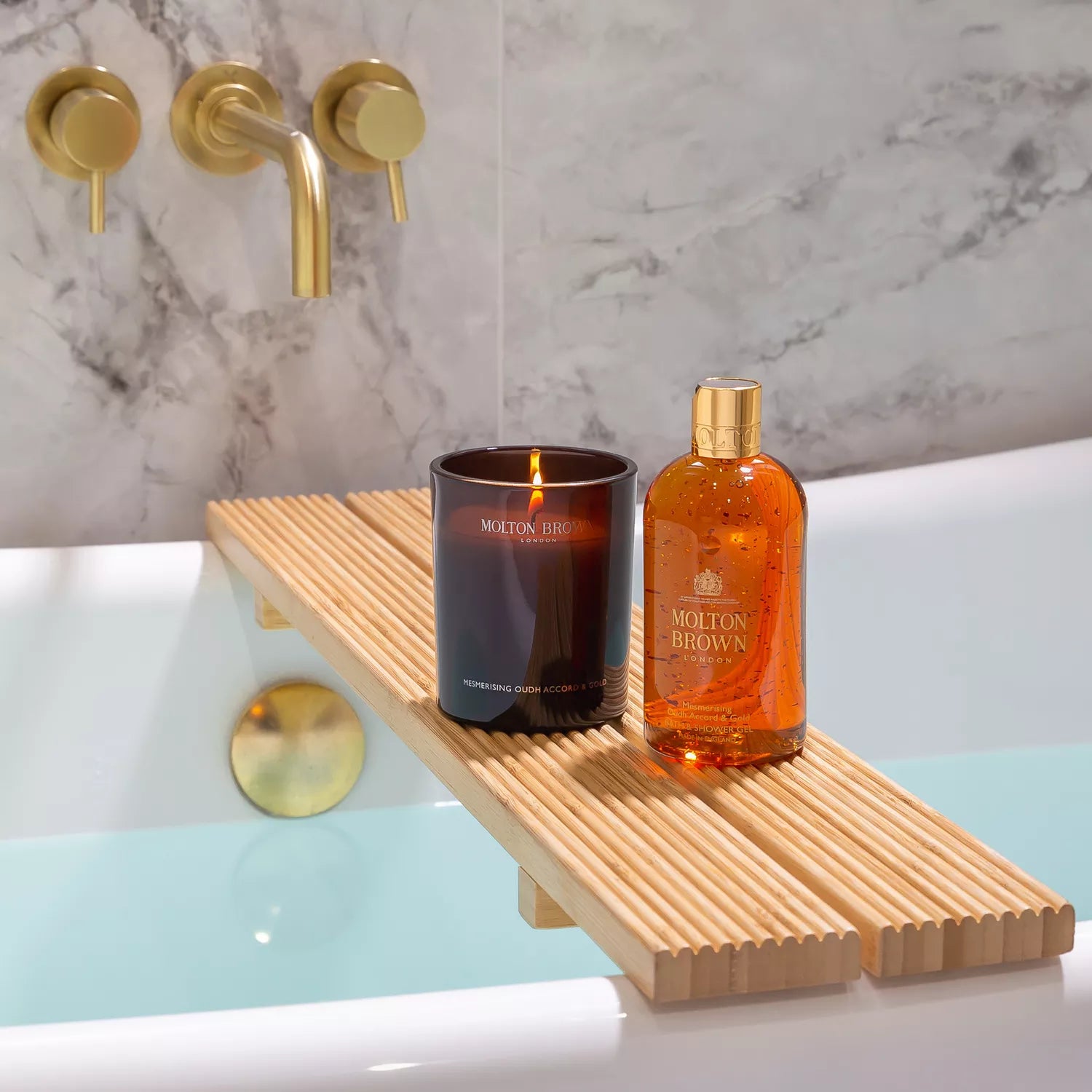 A Mesmerising Oudh Accord & Gold Luxury Kaars 600g by MoltonBrown Nederlands rests on a wooden bath tray over a filled tub, with gold-colored fixtures and marble walls in the background for an elegant oosterse sfeer.