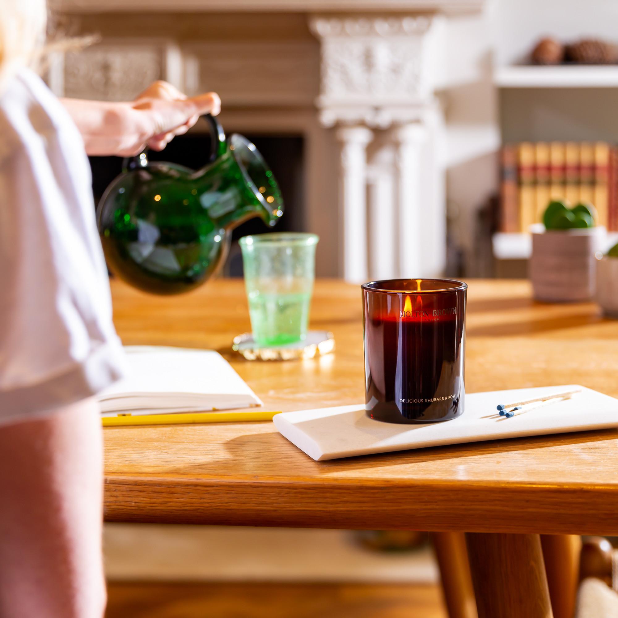 A person pours water from a green glass pitcher into a glass on a wooden table, where a lit MoltonBrown Nederlands Delicious Rhubarb & Rose Signature Kaars 190g, notebook, pen, and small potted plants sit in a cozy, sunlit room.