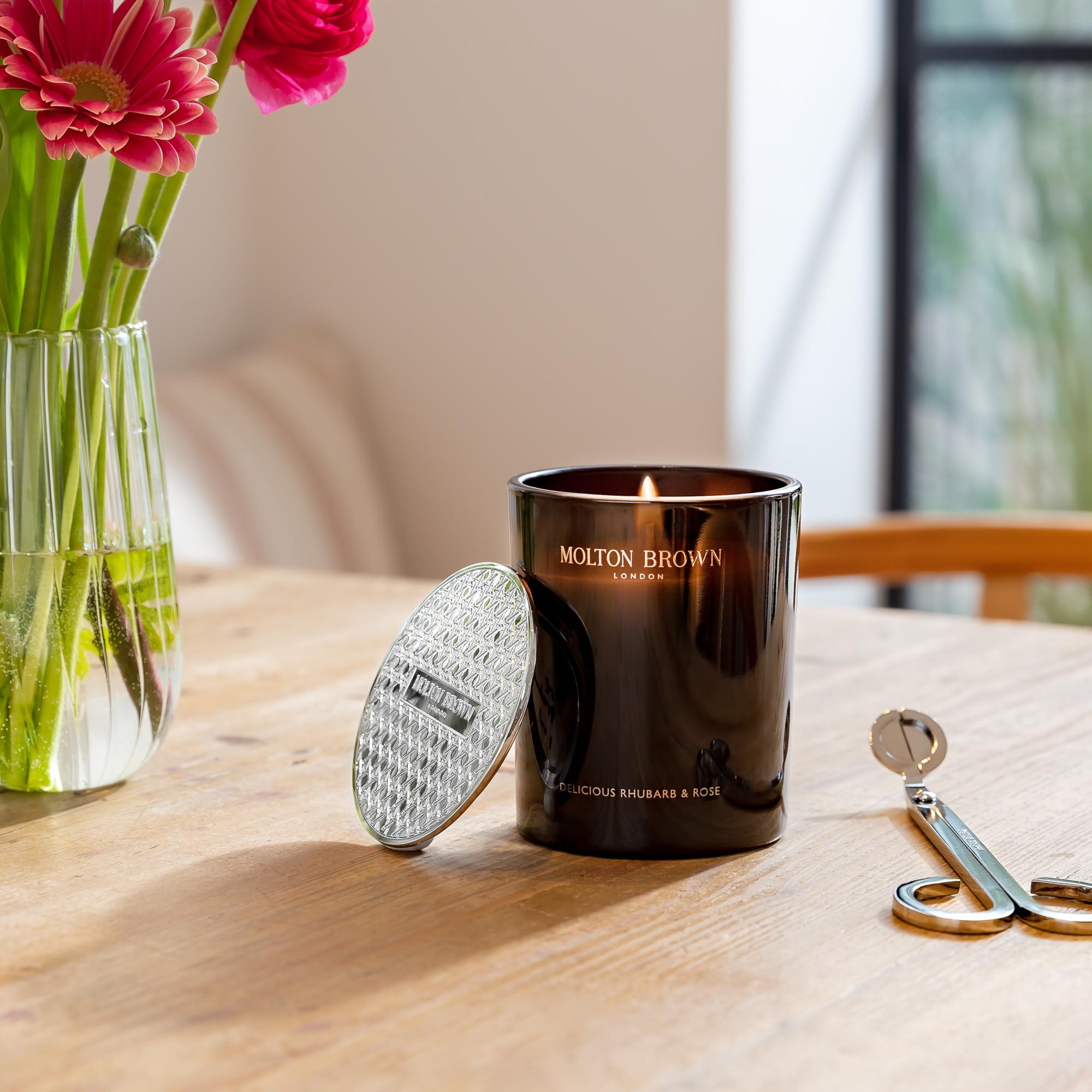A lit MoltonBrown Nederlands Delicious Rhubarb & Rose Signature Kaars 190g sits on a wooden table beside its textured lid, a wick trimmer, and a vase of pink flowers, illuminated by natural light from a nearby window.
