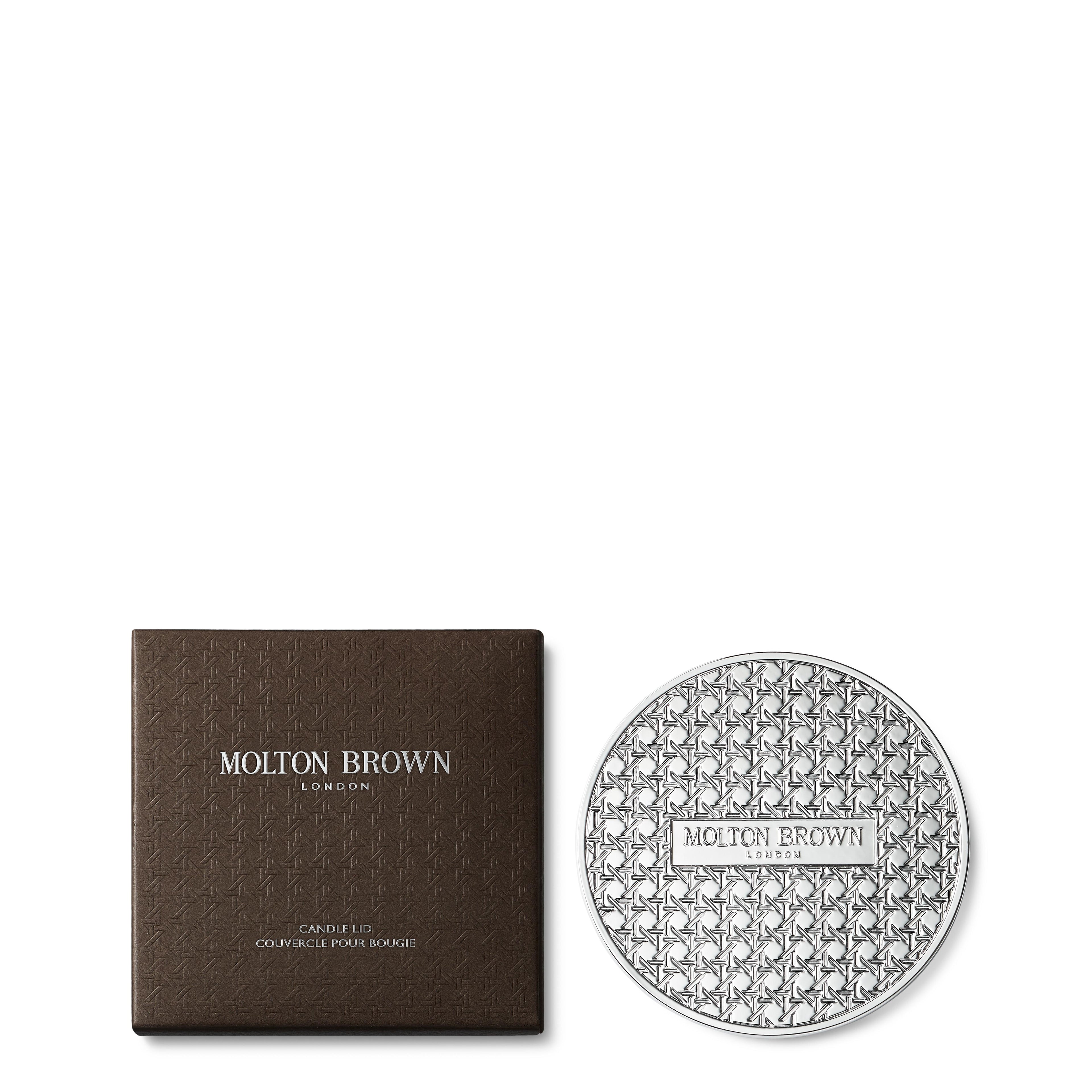 De Signature Kaars Lid 98g van MoltonBrown Nederlands, met zilverkleurig decorpatroon, ligt naast een donkerbruine doos met logo en productinfo—ideaal om je kaars te beschermen en de geur langer te behouden.