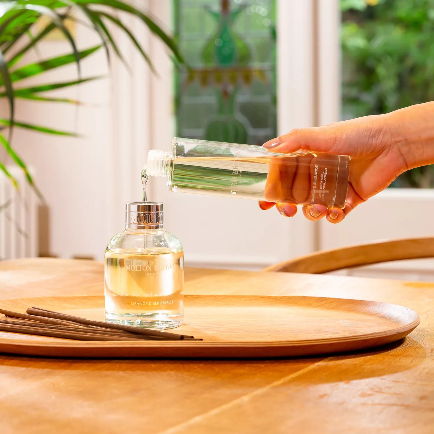 A hand pours liquid from a clear bottle into a glass diffuser on a wooden tray, with MoltonBrown Nederlands Fiery Pink Pepper Perfumed Soap 150g nearby. Sunlight and green plants create a serene indoor atmosphere.