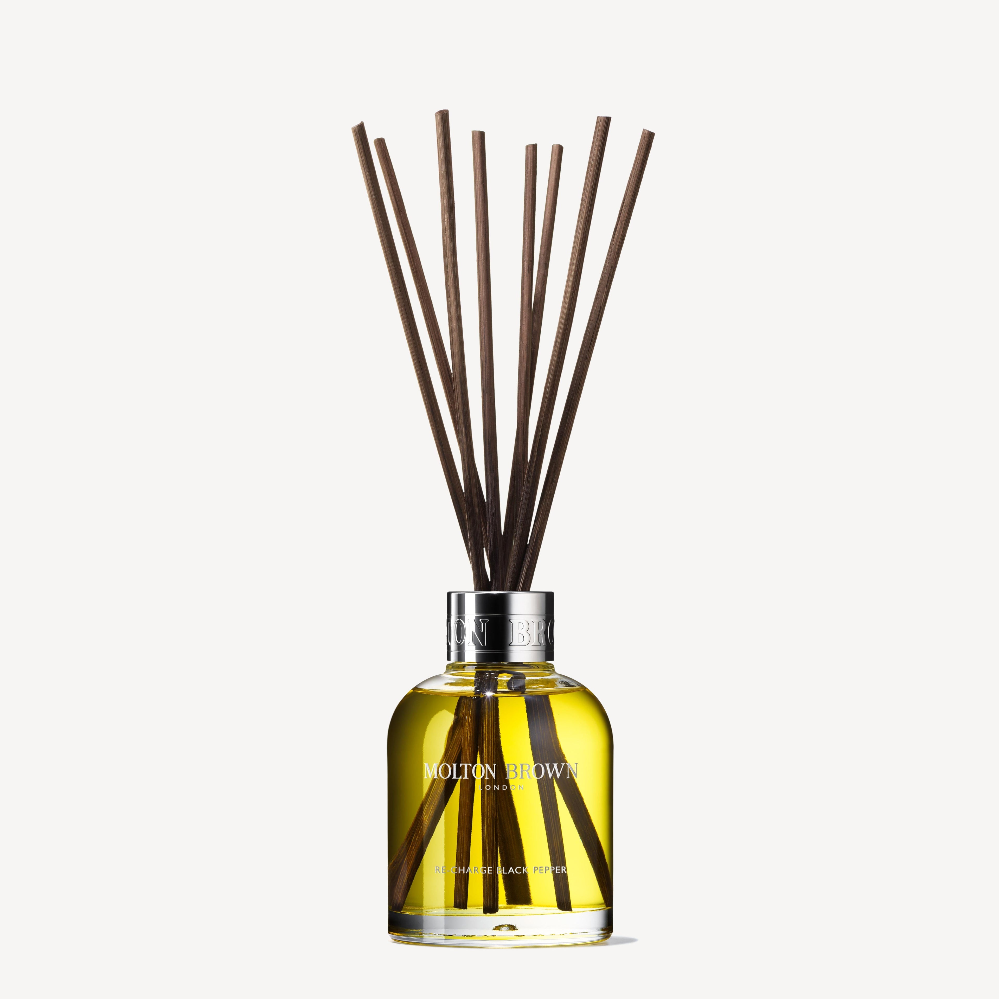 A 150ml clear glass bottle of MoltonBrown Nederlands Re-charge Black Pepper Geurstokjes, with a silver cap and long brown sticks, offers a refined woody-citrus scent.