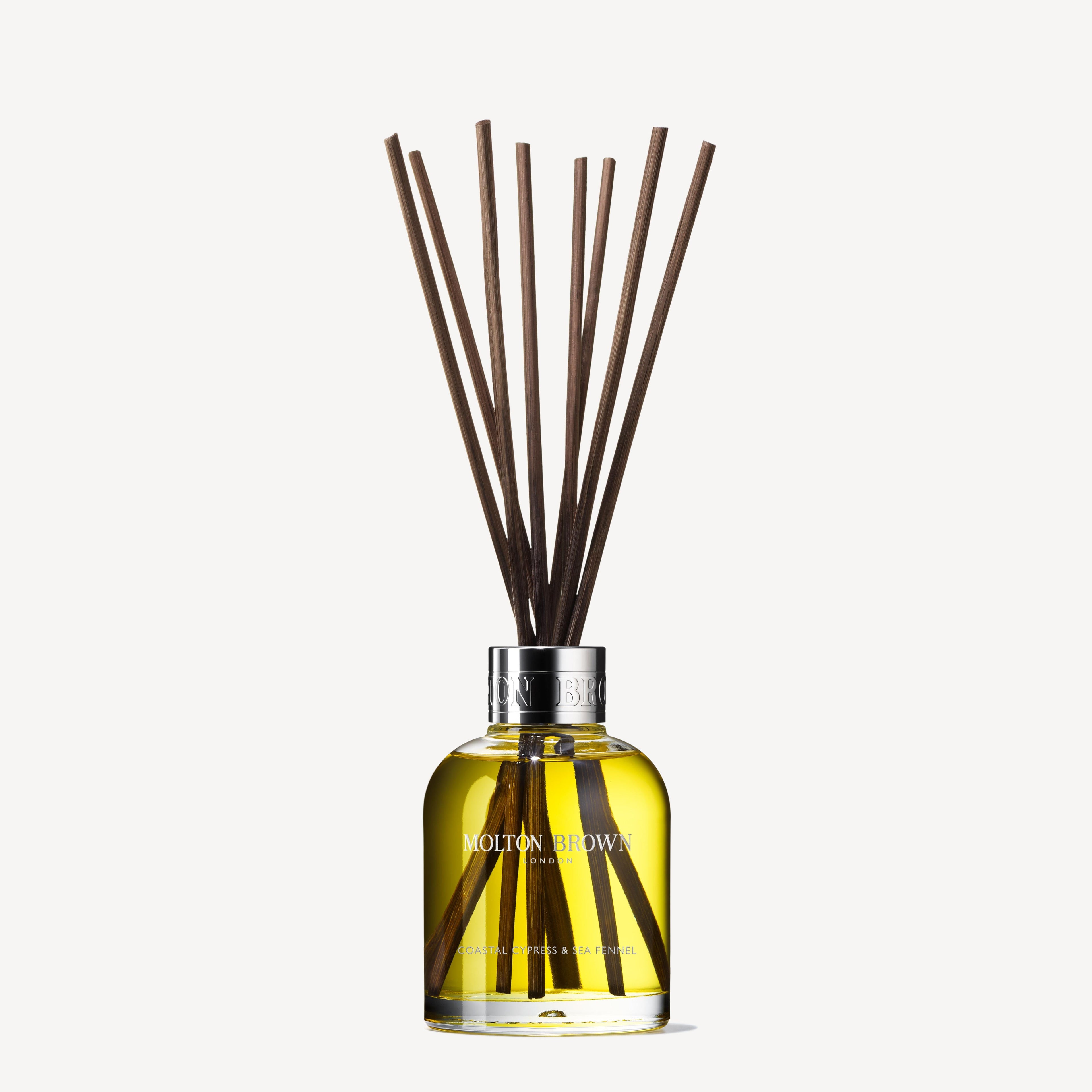 A 150ml glass bottle of MoltonBrown Nederlands Coastal Cypress & Sea Fennel Geurstokjes, filled with yellow liquid and several long, thin scent sticks fanning out to gently release an invigorating coastal fragrance.