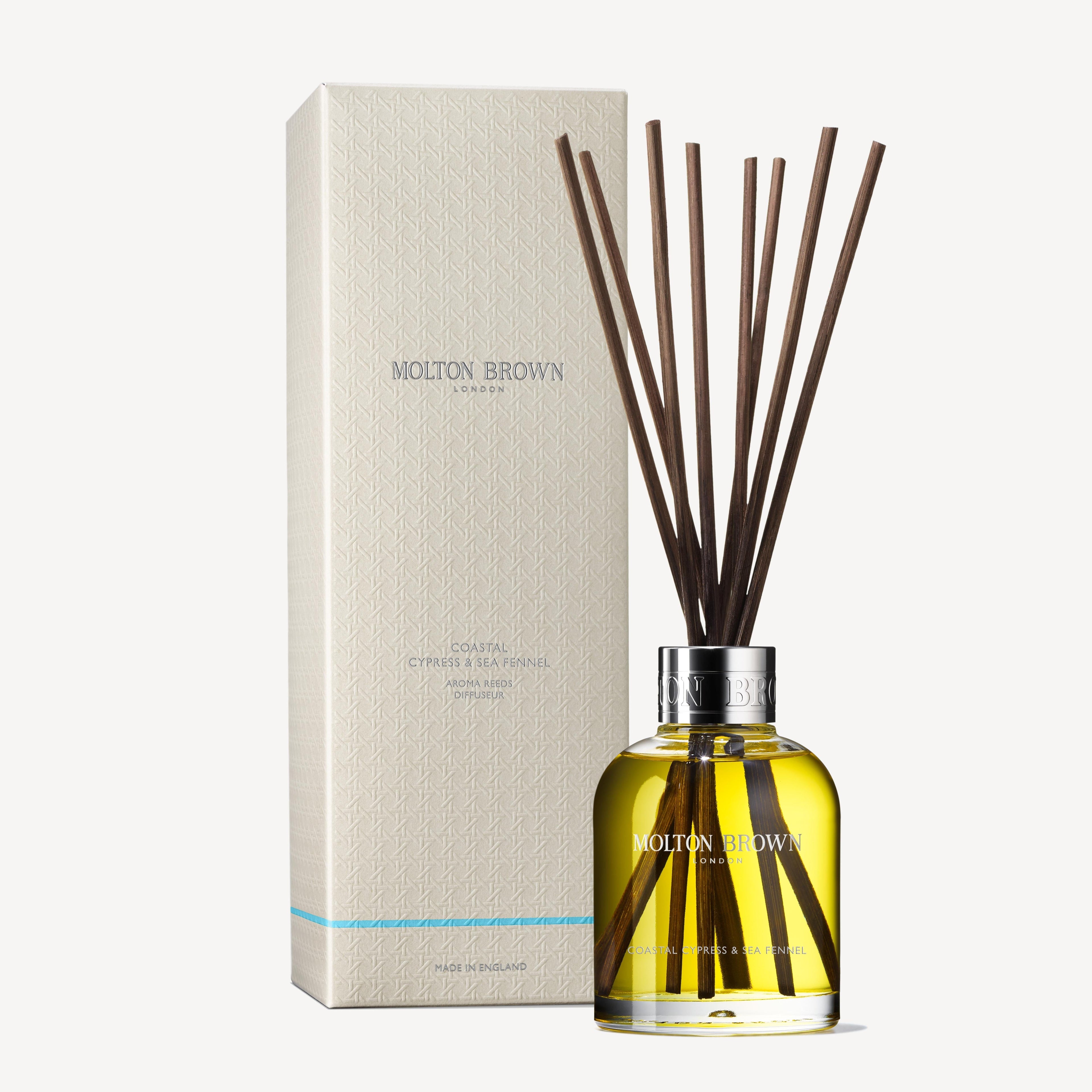 MoltonBrown Nederlands Coastal Cypress & Sea Fennel Geurstokjes 150ml features a glass bottle with yellow liquid and dark sticks, displayed beside its elegant, light-colored box packaging.