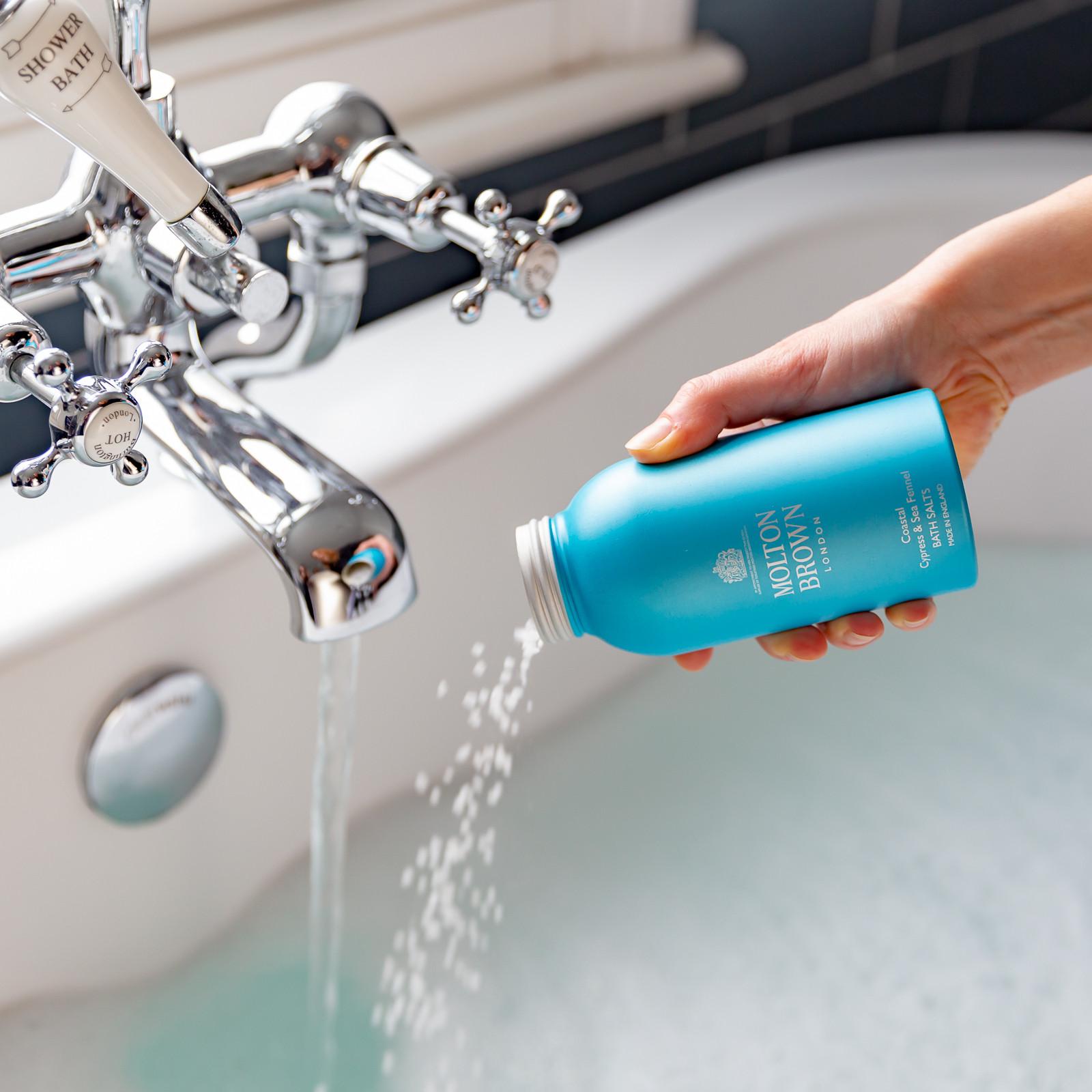 A hand pours Coastal Cypress & Sea Fennel Badzout 300g by MoltonBrown Nederlands from a blue bottle into a bathtub with running water beneath a silver faucet.