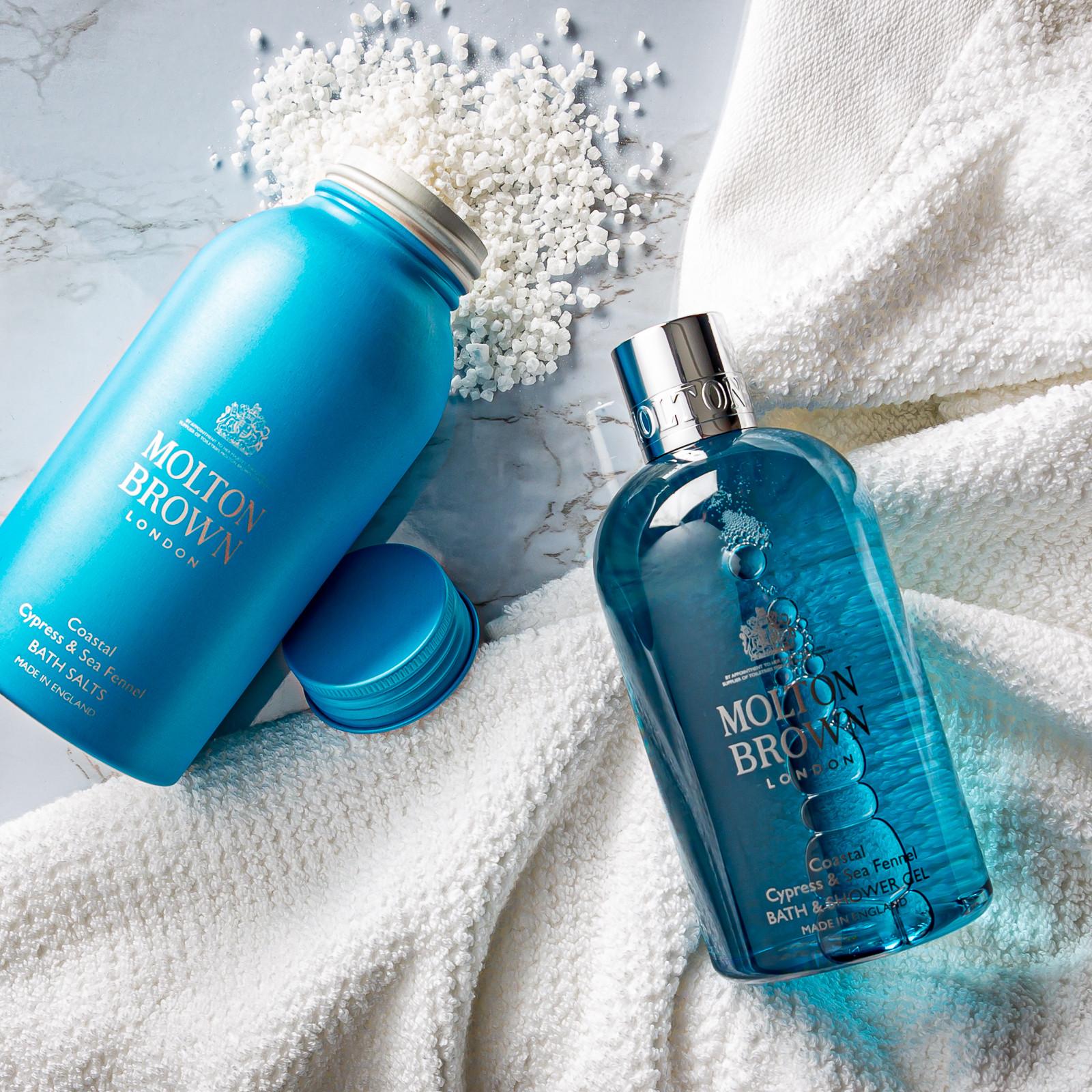MoltonBrown Nederlands Coastal Cypress & Sea Fennel Badzout 300g and a handzeep with warme musk in blue bottles rest on a white towel; one bottle is open, with bath salts scattered across the marble surface.