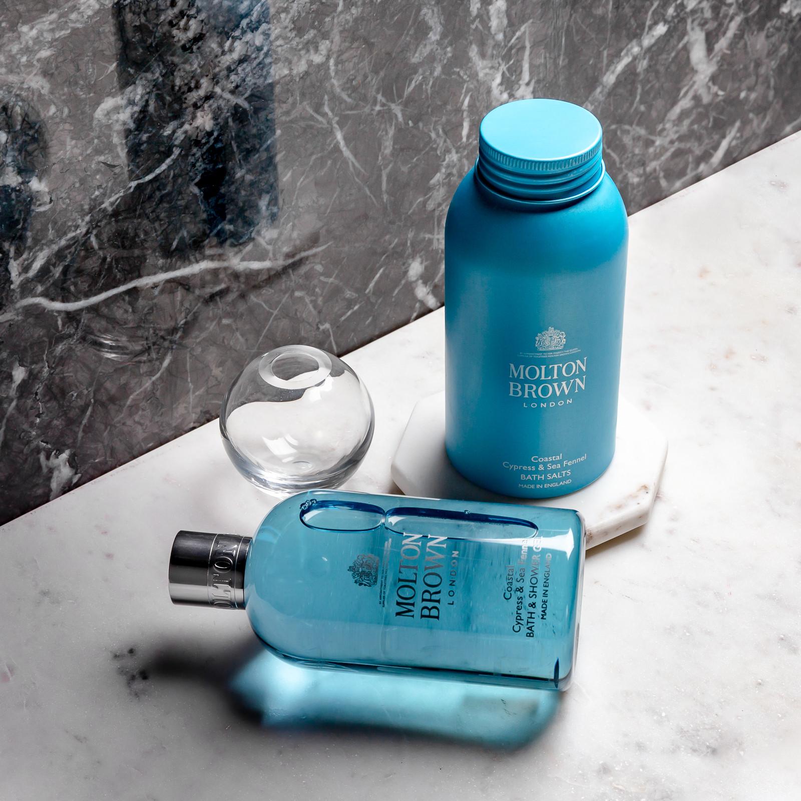 Two blue MoltonBrown Nederlands bath products, including Coastal Cypress & Sea Fennel Badzout 300g, are displayed on a marble surface beside a small clear glass object against a grey marble background.