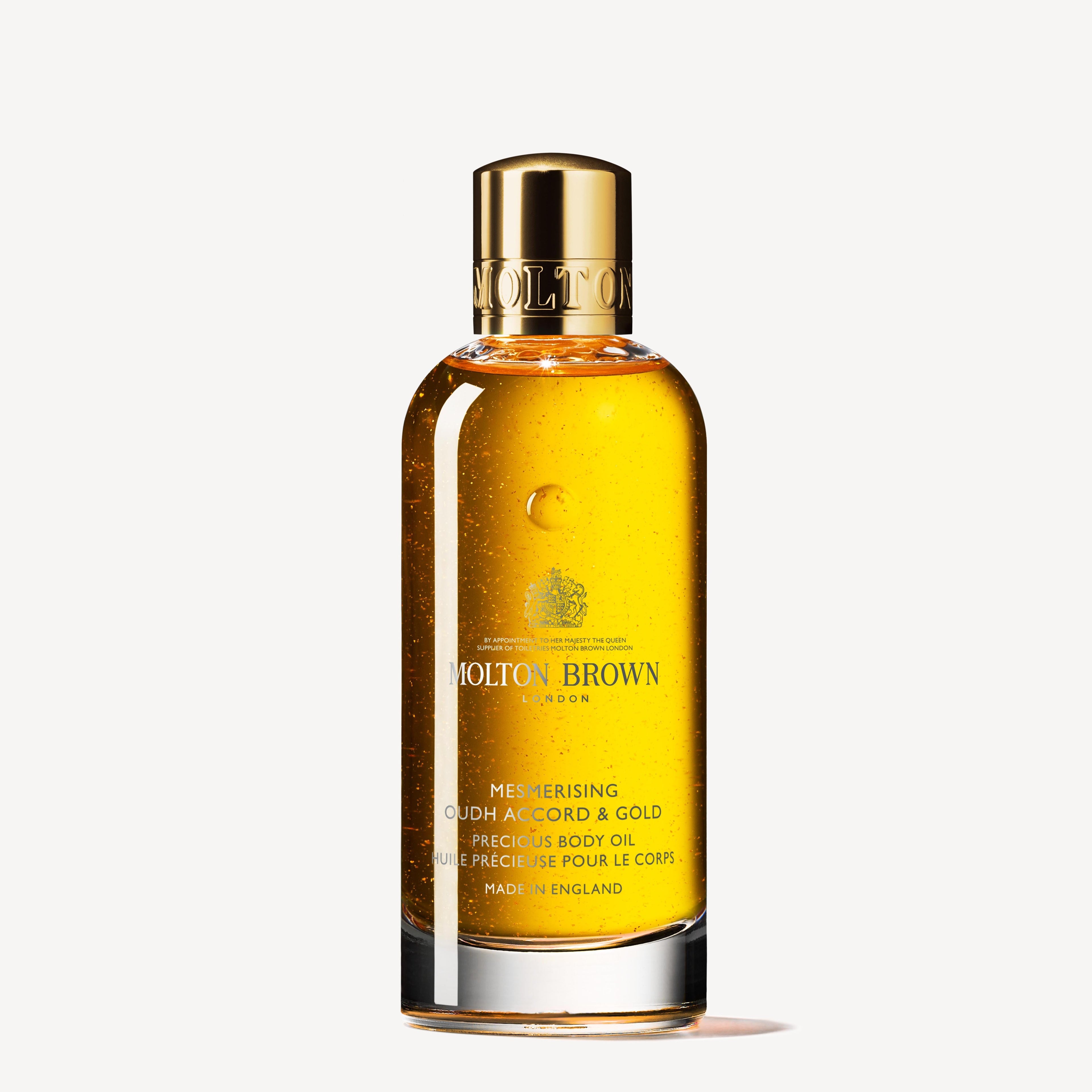 A clear bottle of MoltonBrown Nederlands Mesmerising Oudh Accord & Gold Precious Lichaamsolie 100ml, with a gold cap and label, contains golden oil with small gold flakes, shown against a plain white background.