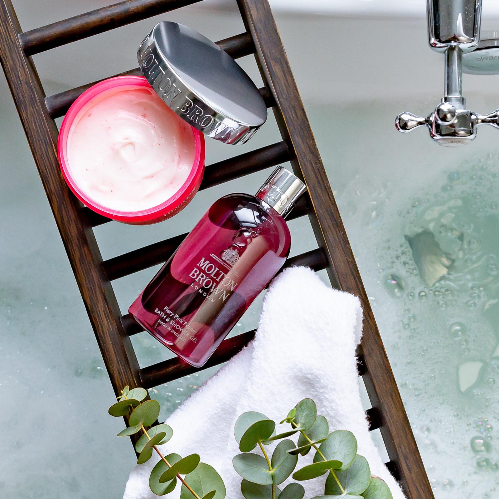 A jar of MoltonBrown Nederlands Fiery Pink Pepper Pampering Body Polisher 275g, a white towel, and eucalyptus leaves sit on a dark wooden tray above a frothy bathtub.