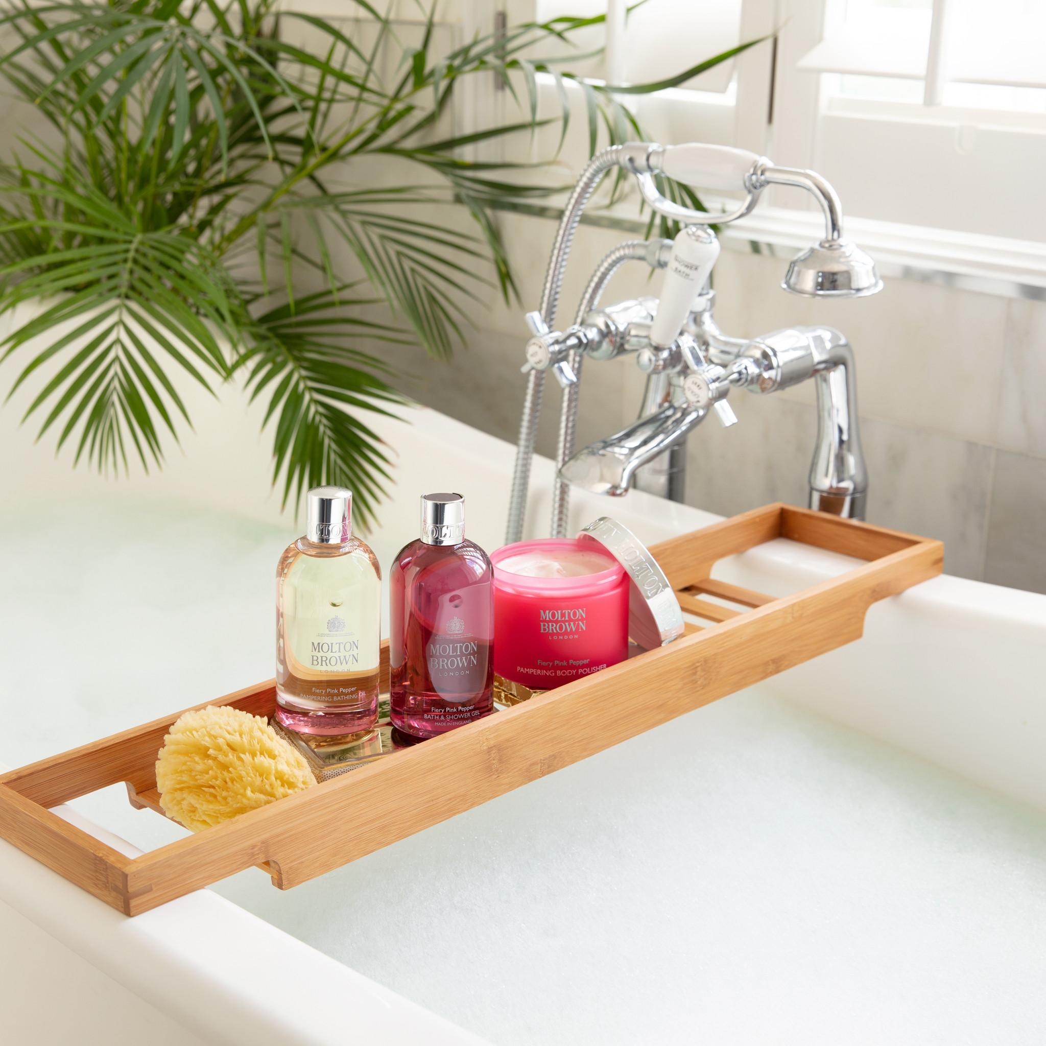A bamboo bath tray holds MoltonBrown Nederlands Fiery Pink Pepper Pampering Body Polisher 275g, a sponge, and a pink candle above a bubble-filled tub, with a chrome faucet and leafy green plant in the background.