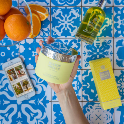 A hand holds a jar of MoltonBrown Nederlands Orange & Bergamot Radiant Body Polisher 275g above blue and white patterned tiles, with oranges, a Molton Brown bottle, a phone, and a yellow box also in view.