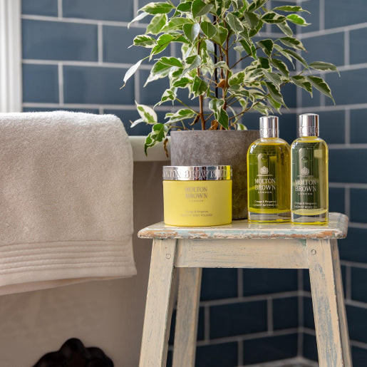 A rustic wooden stool holds a potted plant, a jar, and two MoltonBrown Nederlands Orange & Bergamot Radiant Body Polisher 275g jars next to a bathtub with white towels, set against dark blue subway tile walls.