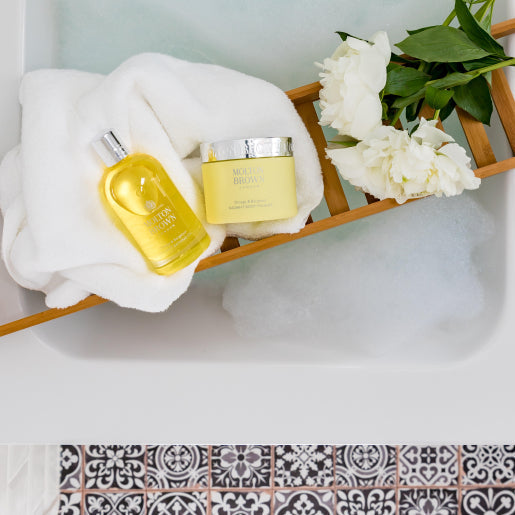 A wooden bath tray holds white towels, MoltonBrown Nederlands Orange & Bergamot Radiant Body Polisher 275g, and white flowers above a bubble-filled bathtub with black-and-white patterned floor tiles visible below.