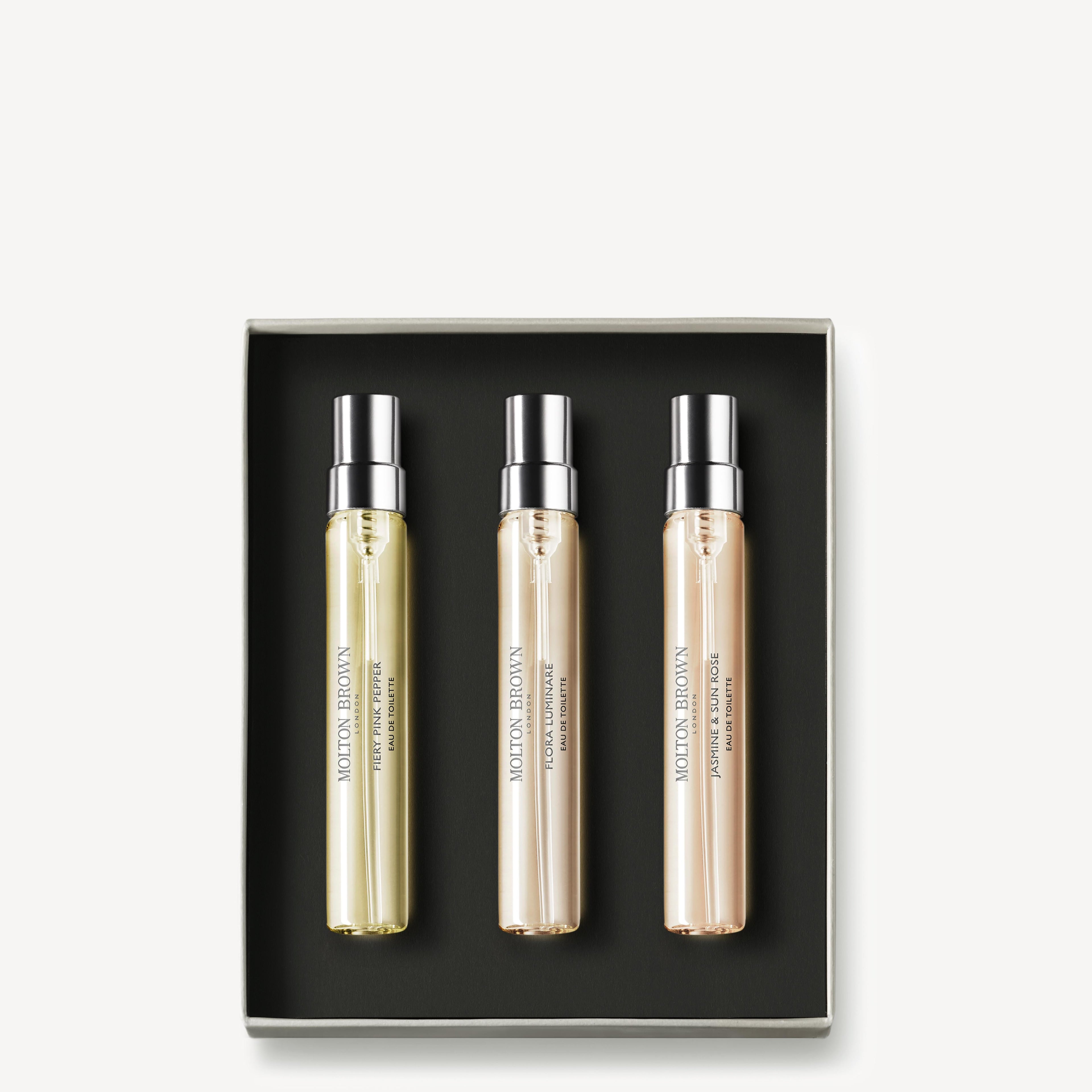 A square box from MoltonBrown Nederlands contains three Floral & Spicy Fragrance Ontdekkingsset bottles—pale yellow, gold, and light peach—neatly arranged side by side on a black background.