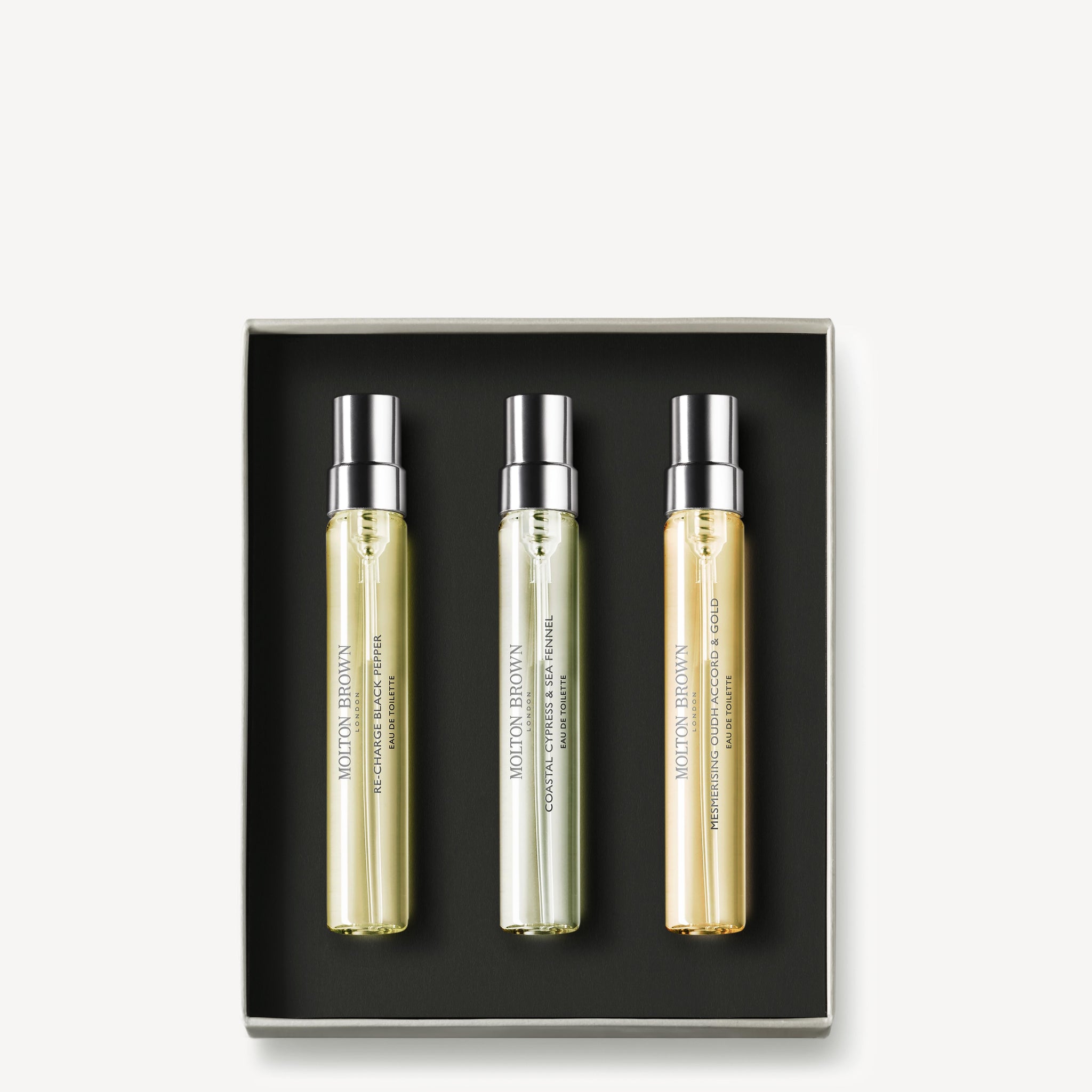 A box of MoltonBrown Nederlands' Aromatic & Ambery Fragrance Ontdekkingsset features three clear glass bottles with metallic caps, each labeled with a unique scent, arranged side by side on a black background.